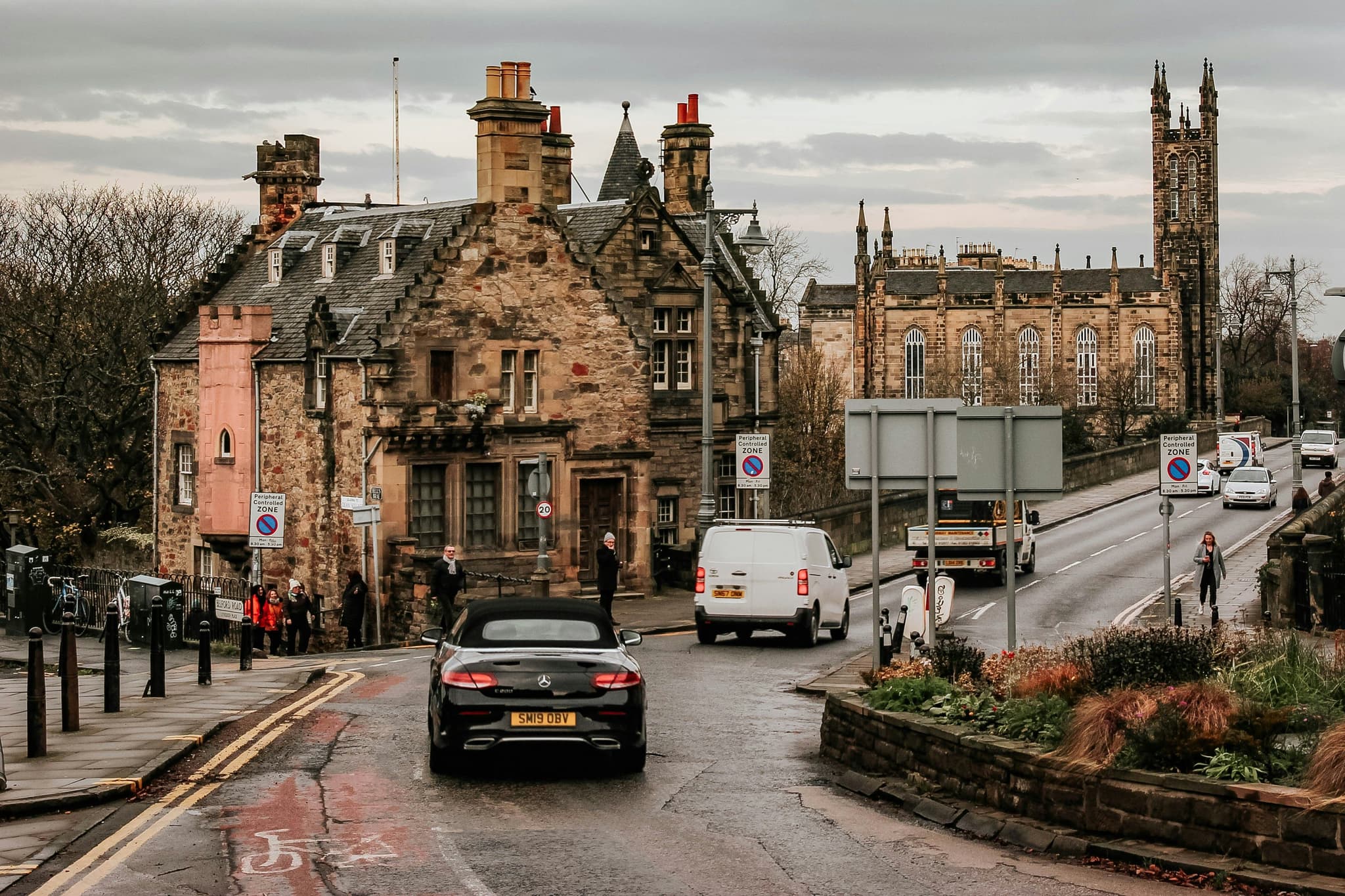 Cars driving on a road in Scotland next to historic buildings and churches on a rainy day.
