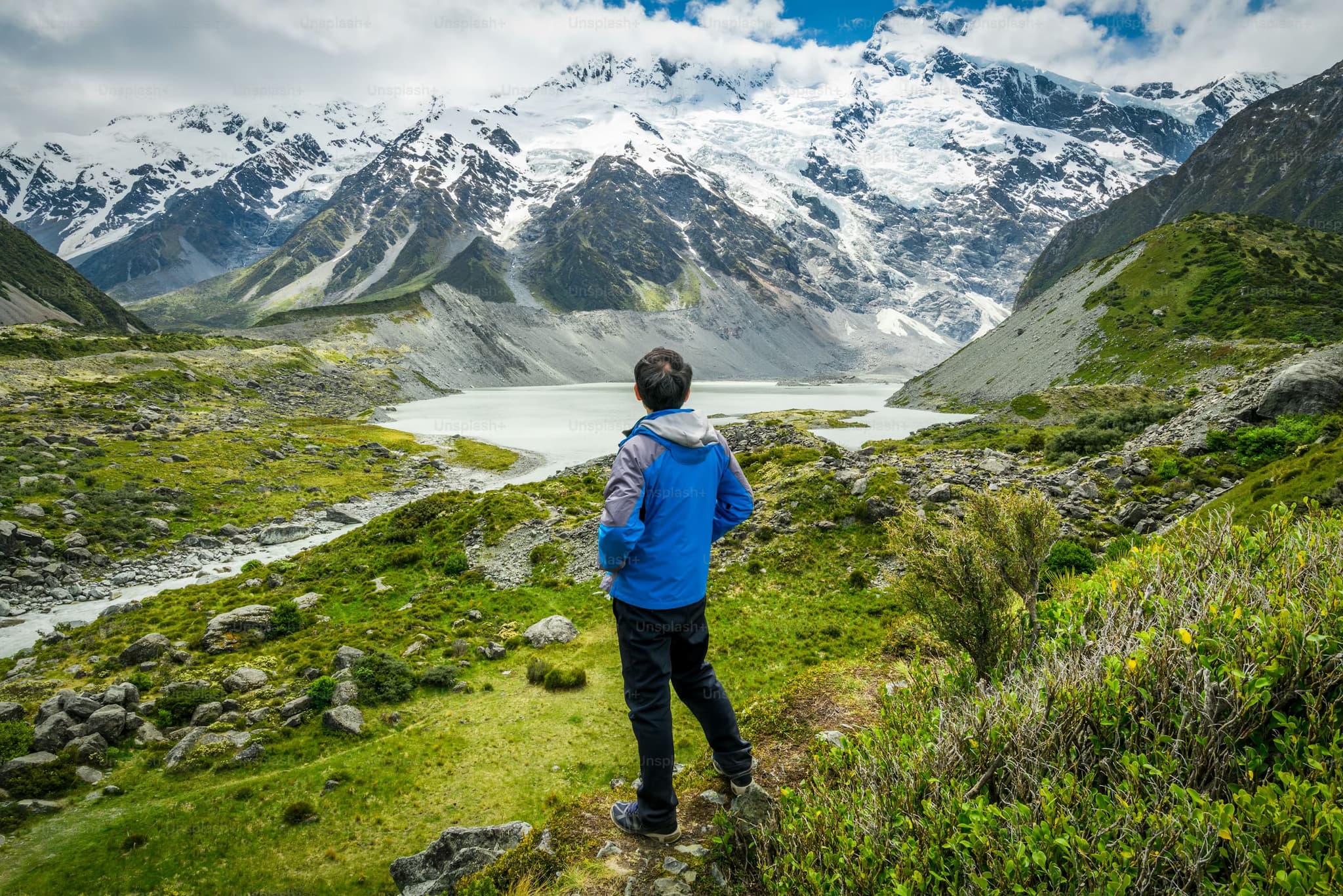 A person in a blue jacket stands on a grassy hill, overlooking a serene mountain landscape with snow-capped peaks and a tranquil lake, evoking a sense of contemplation.