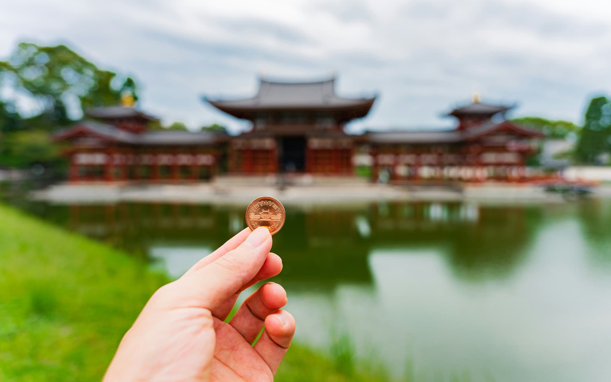 A hand holding a coin in front of a lake with japanese red temple