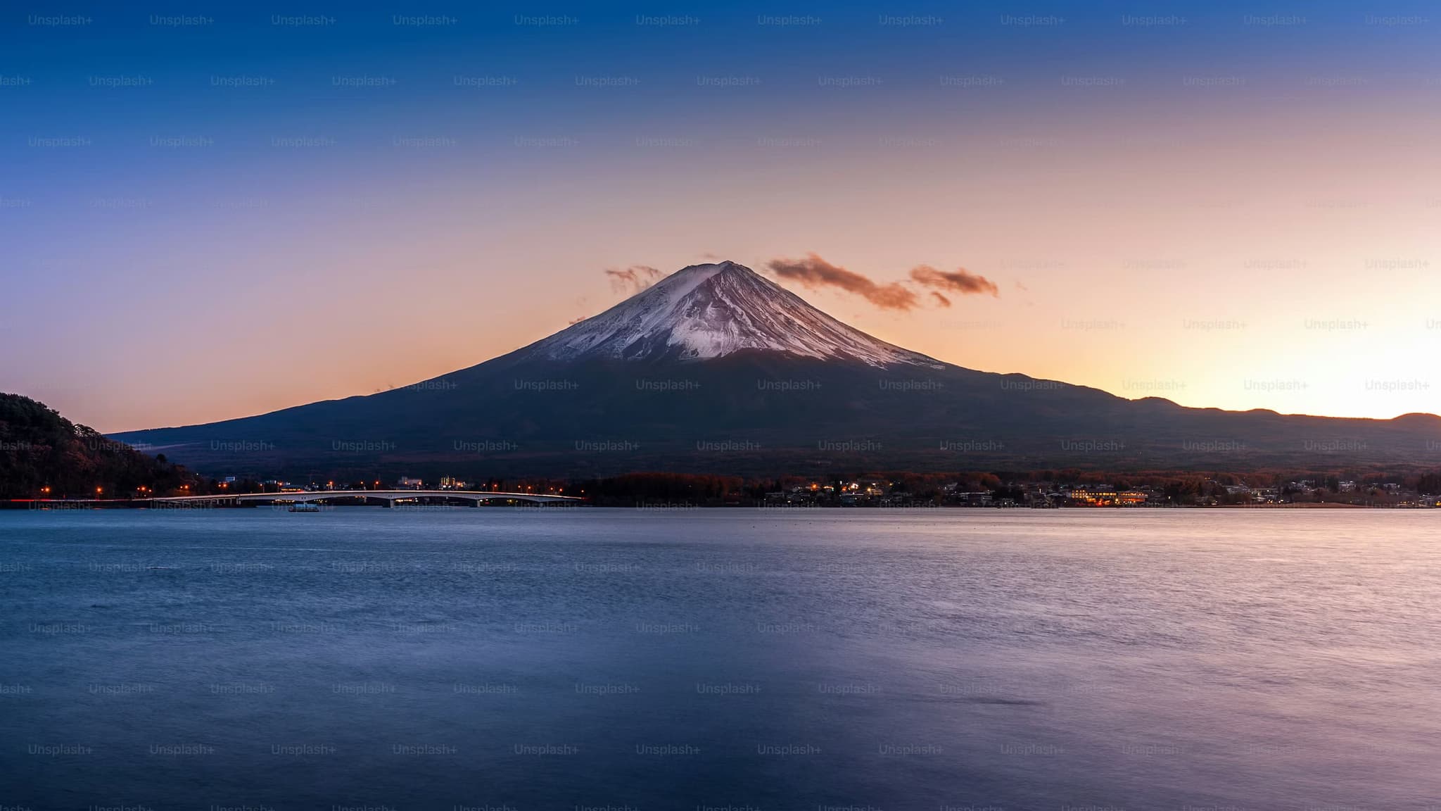 Sunset over Mount Fuji with a snow-capped peak, reflected in calm waters. The sky transitions from warm orange to cool blue, creating a tranquil scene.