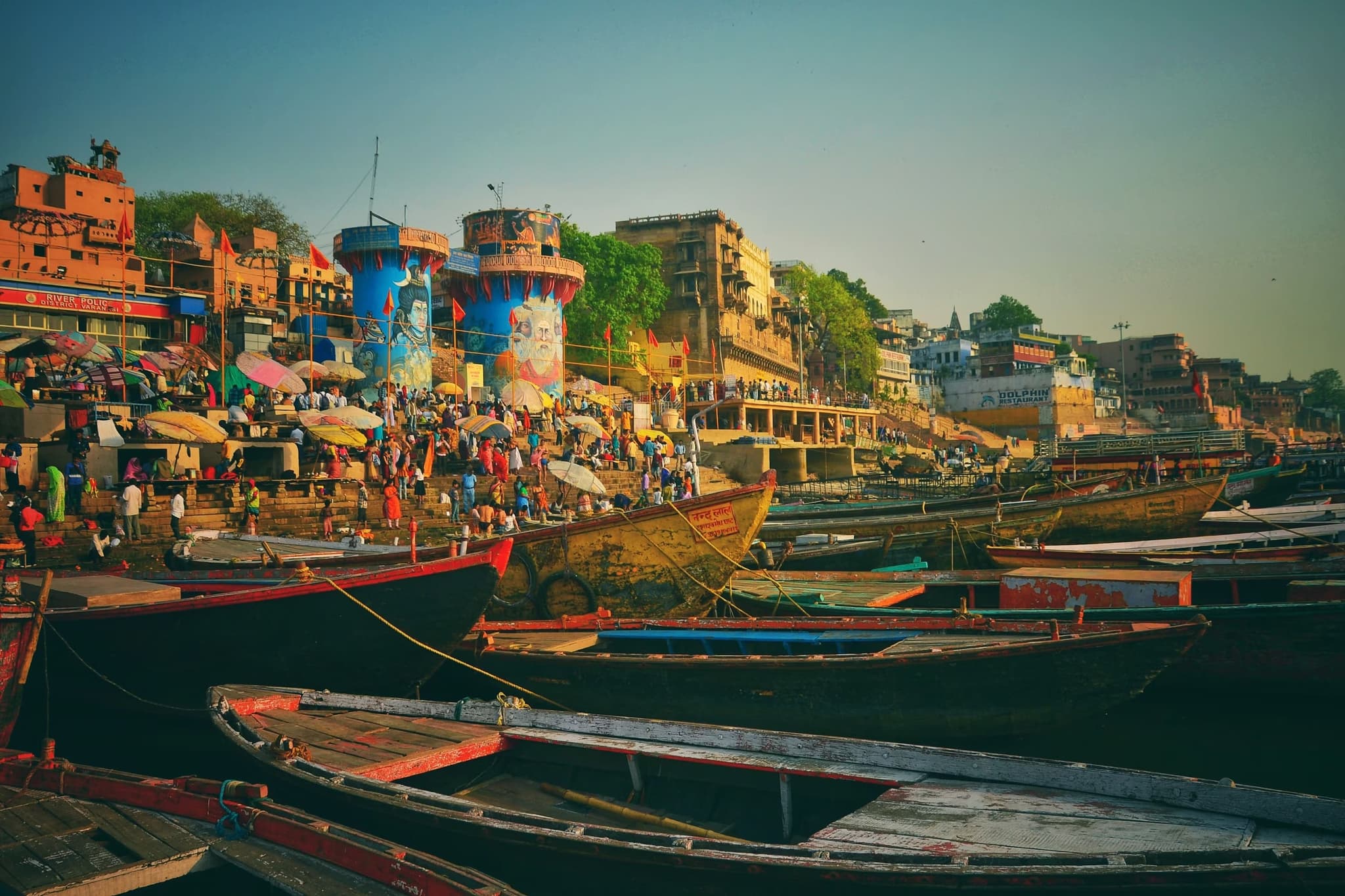 A traditional houseboat floats on a calm river surrounded by lush palm trees under a clear blue sky, creating a serene and picturesque setting.