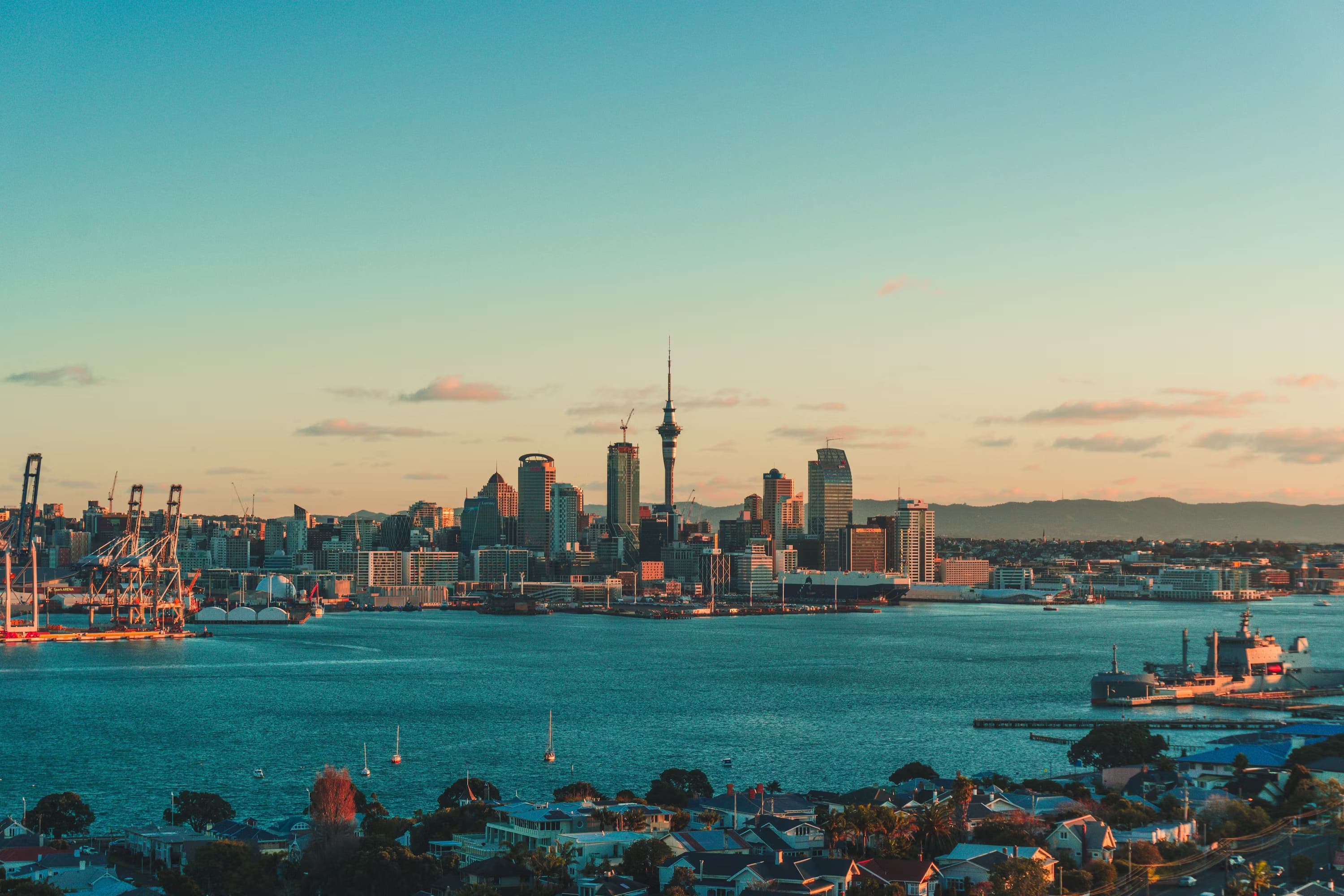 Auckland city skyline at sunset, featuring the Sky Tower, silhouetted against a warm sky. The harbor in the foreground has calm blue waters.