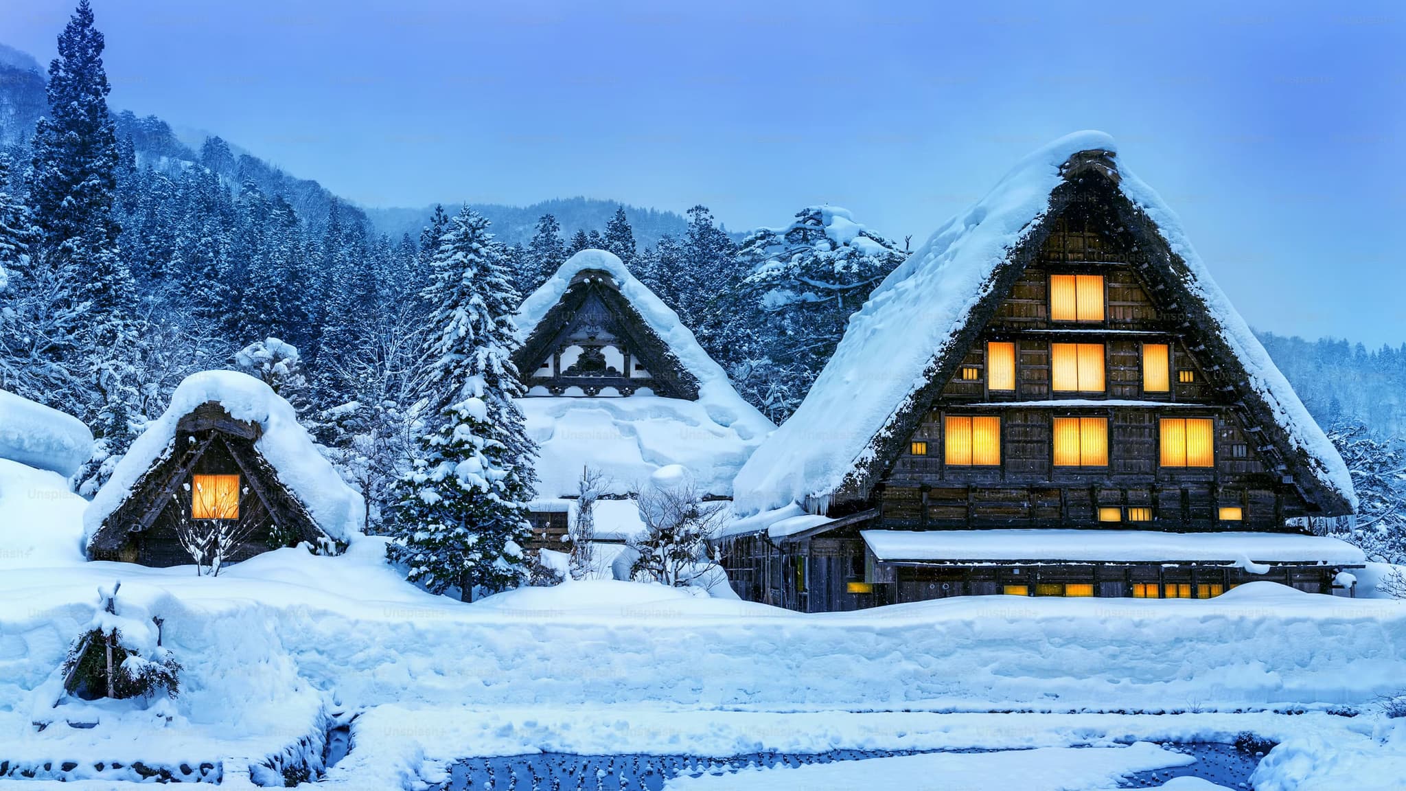 Traditional wooden houses with steep, snow-covered thatched roofs glow warmly in a serene, snowy landscape with fir trees and hills at dusk.