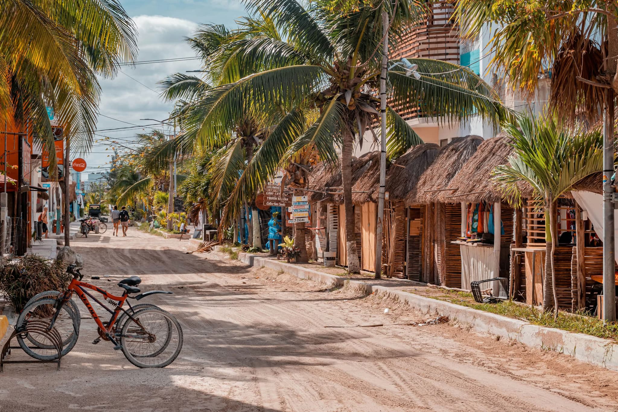 A sandy street lined with palm trees and rustic wooden huts under a bright blue sky. A red bicycle rests on the left, creating a laid-back, tropical vibe.