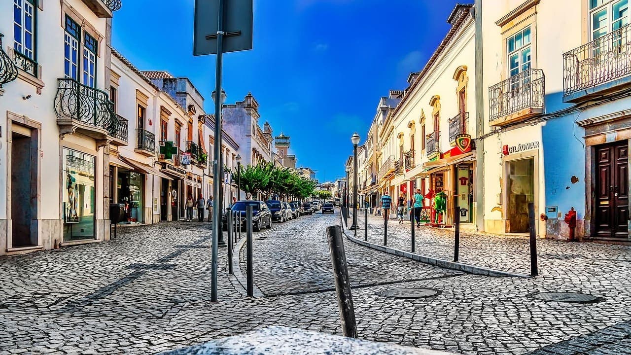 A colourful cobbled street in Portugal with pastel buildings, small shops and balconies under a bright blue sky.