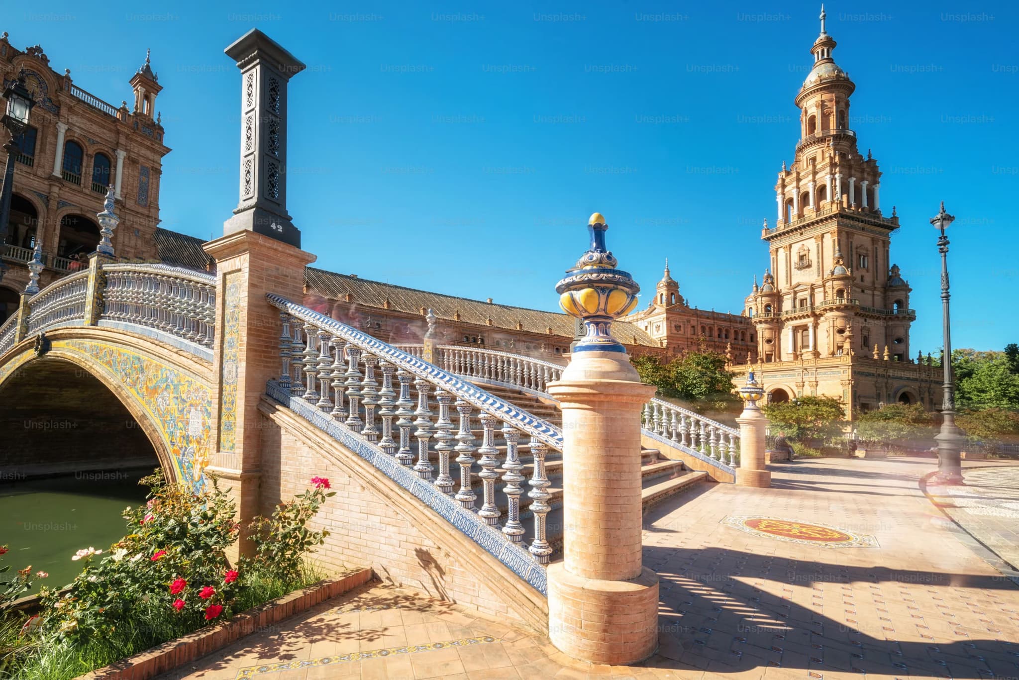 Ornate arched bridge with ceramic tiles in Plaza de España, Seville. Sunlit tower and vibrant blue sky evoke a sense of historic elegance and warmth.