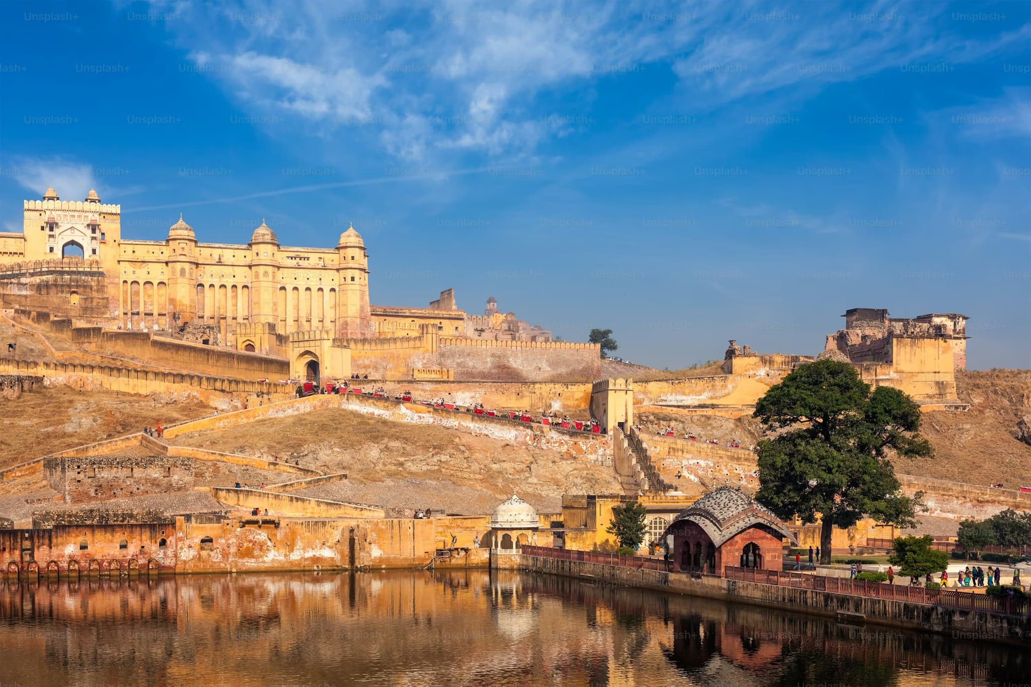 Historic Amber Fort under a vibrant blue sky, with beige sandstone walls, lush greenery, and a reflective water body in the foreground.