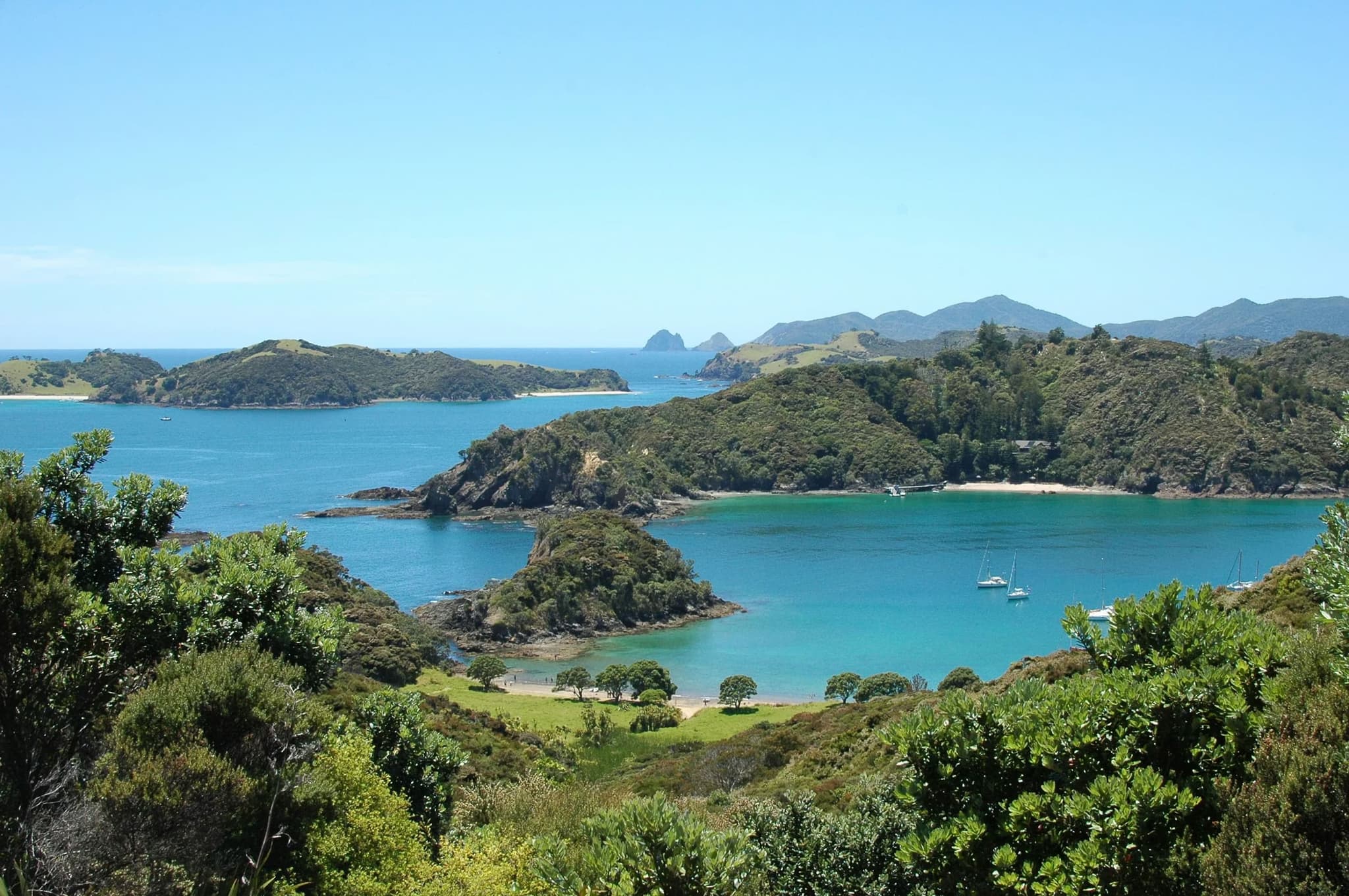 Scenic coastal view of lush green islands and turquoise waters under a clear blue sky. A few sailboats are anchored in the tranquil bay.