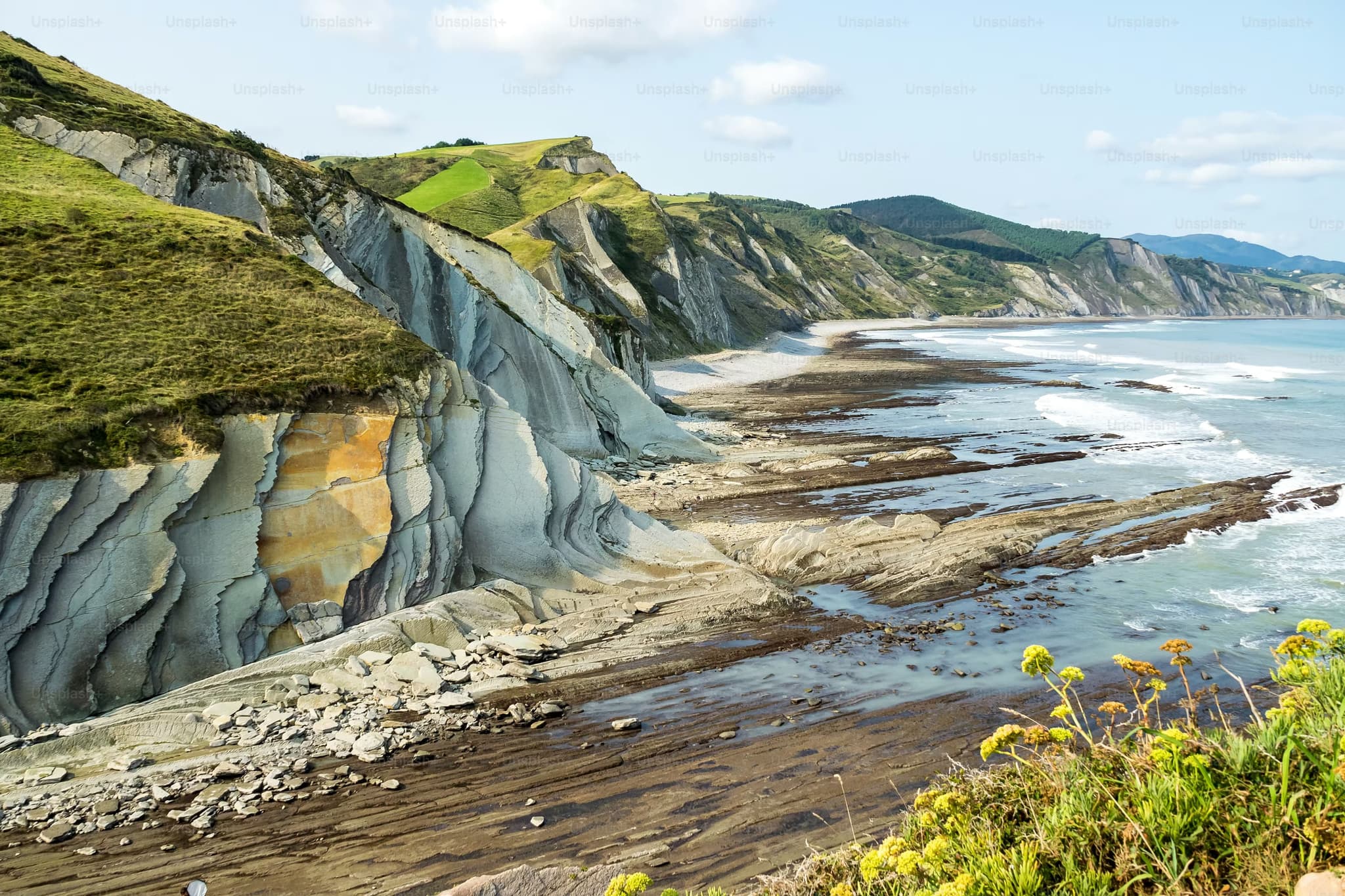 Rugged coastal cliffs with striated rock formations, lush green slopes, and a narrow beach meet the ocean's edge under a partly cloudy sky.
