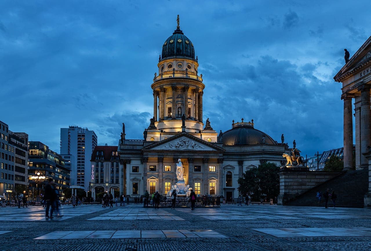 Berlin Cathedral illuminated at dusk, with people walking across the large square in front of Museum Island.