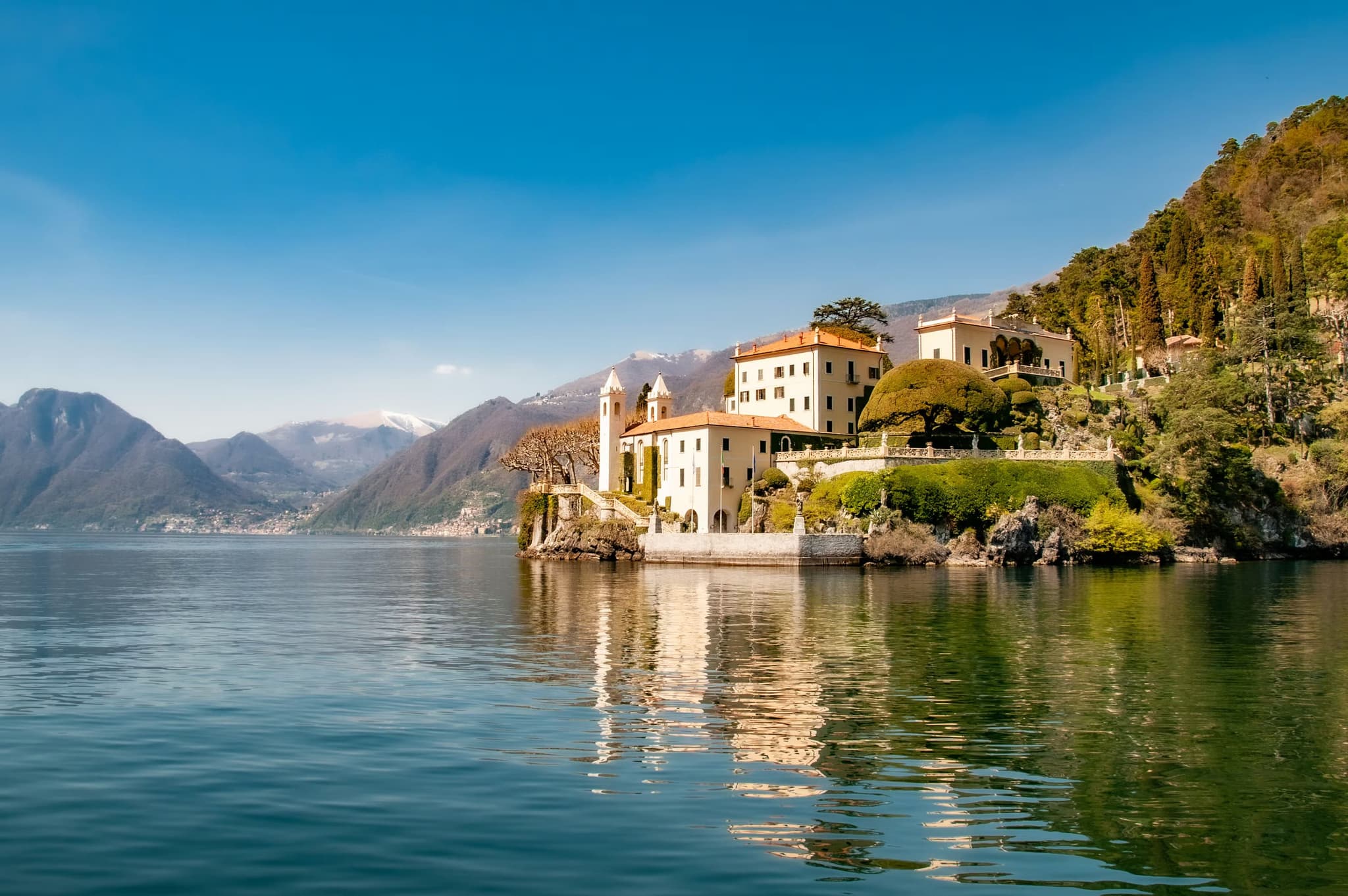 Scenic view of a villa on Lake Como's edge, flanked by lush greenery and mountains. Calm water reflects the clear blue sky and elegant architecture.