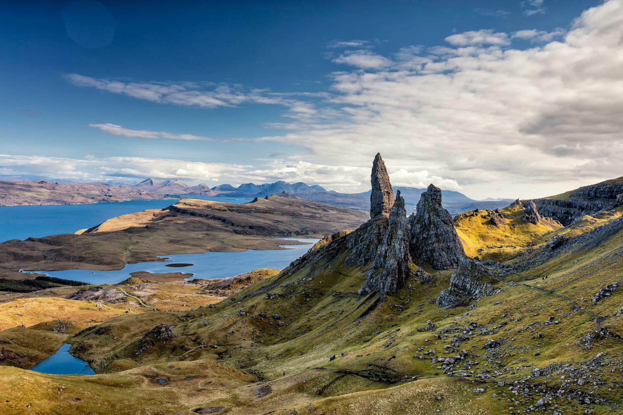 Aerial view of Isle of Skye: green landscape with high mountain rock peaks and lake in the distance on a cloudy day with blue sky
