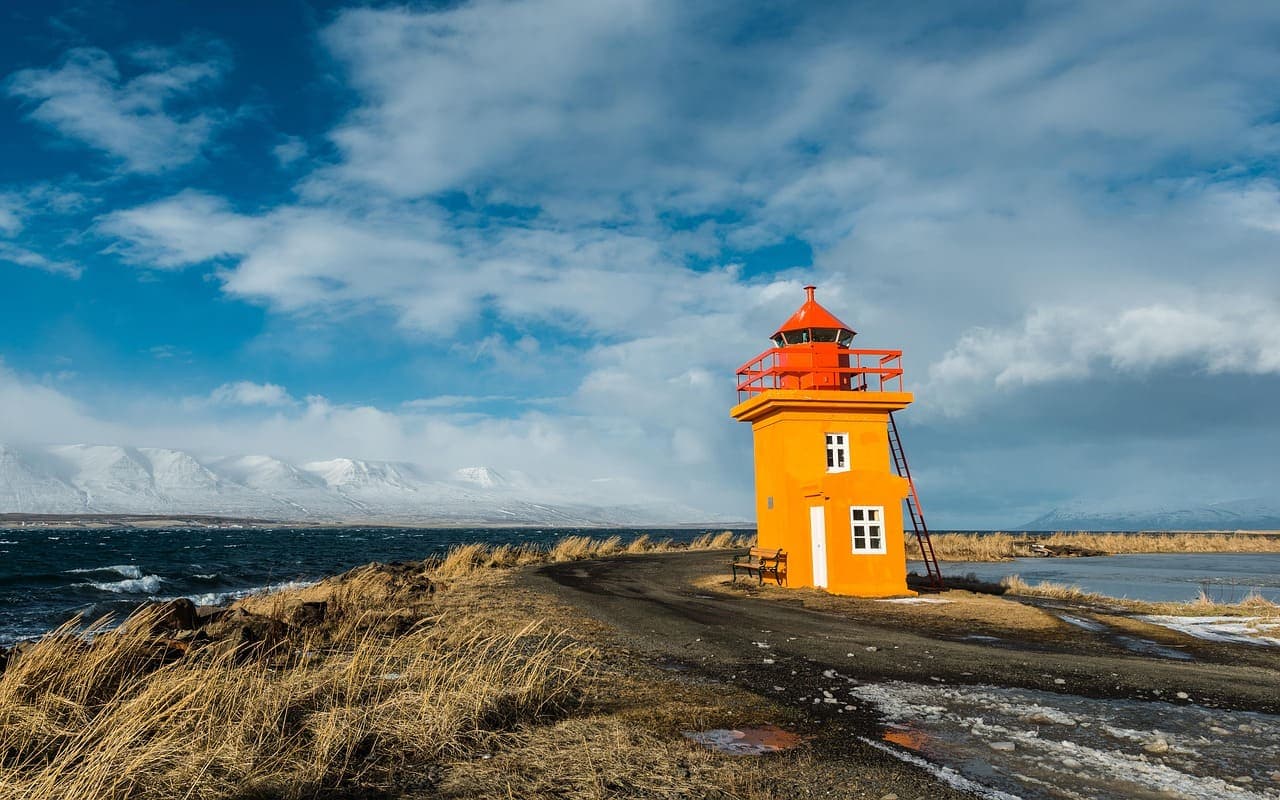 Bright yellow lighthouse on a rocky Icelandic coastline with snowy mountains and a blue sky in the background.