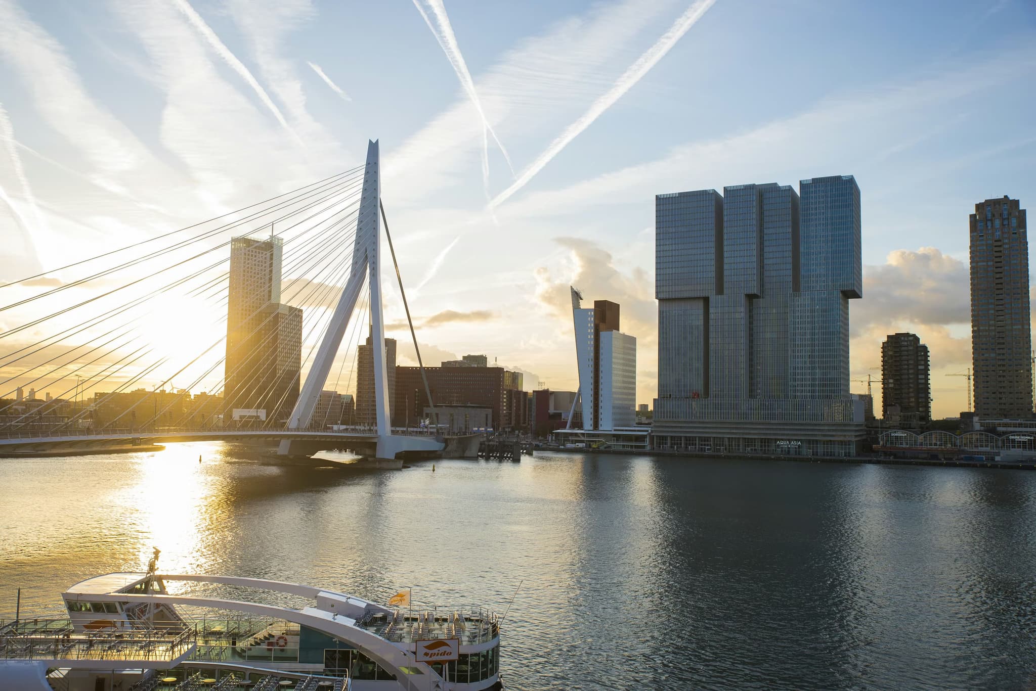 Sunset view of Rotterdam's skyline with the Erasmus Bridge in the foreground, modern skyscrapers reflecting on the calm river under a pastel sky.