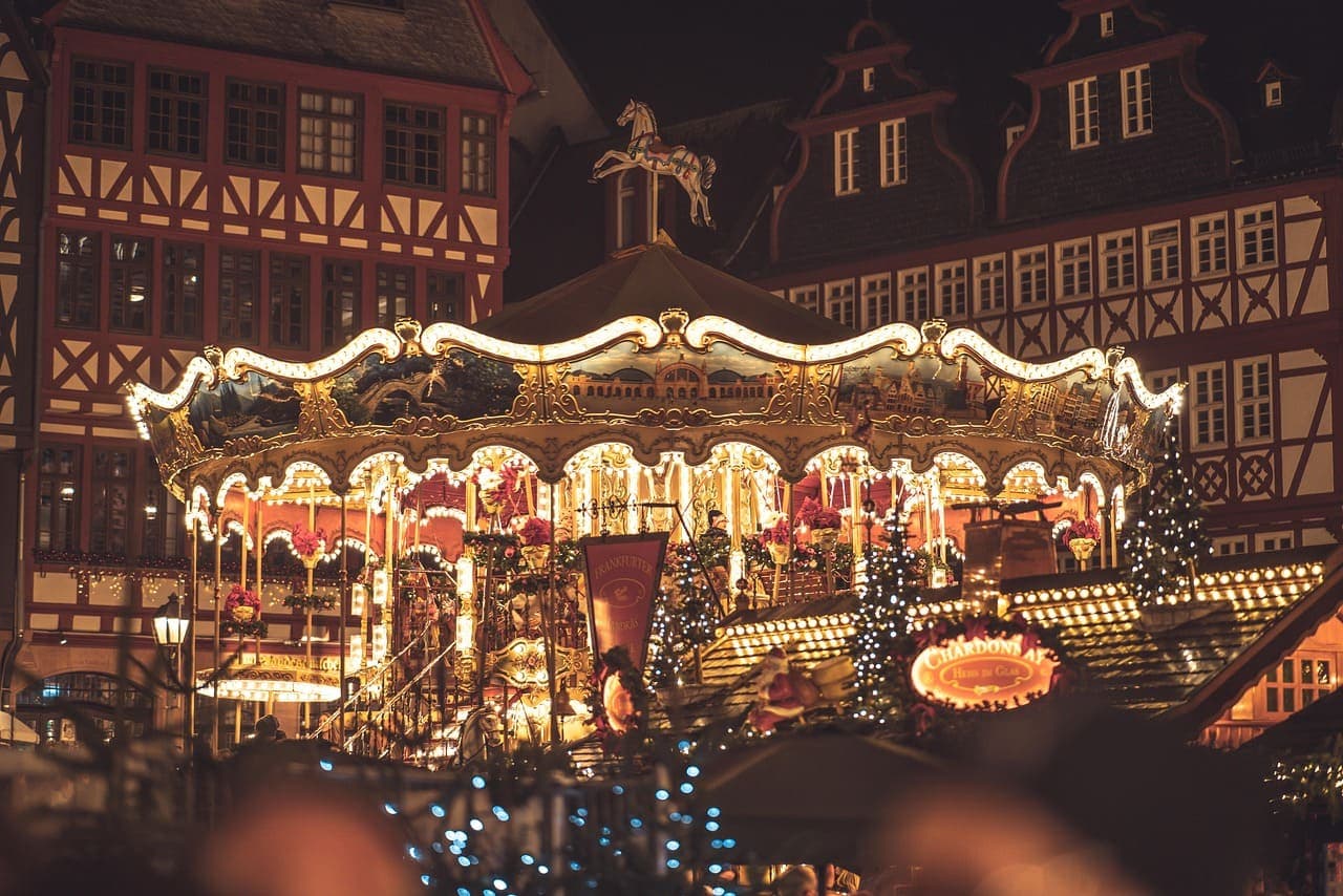 Illuminated Christmas market carousel at night with festive lights and traditional European style buildings in the background.