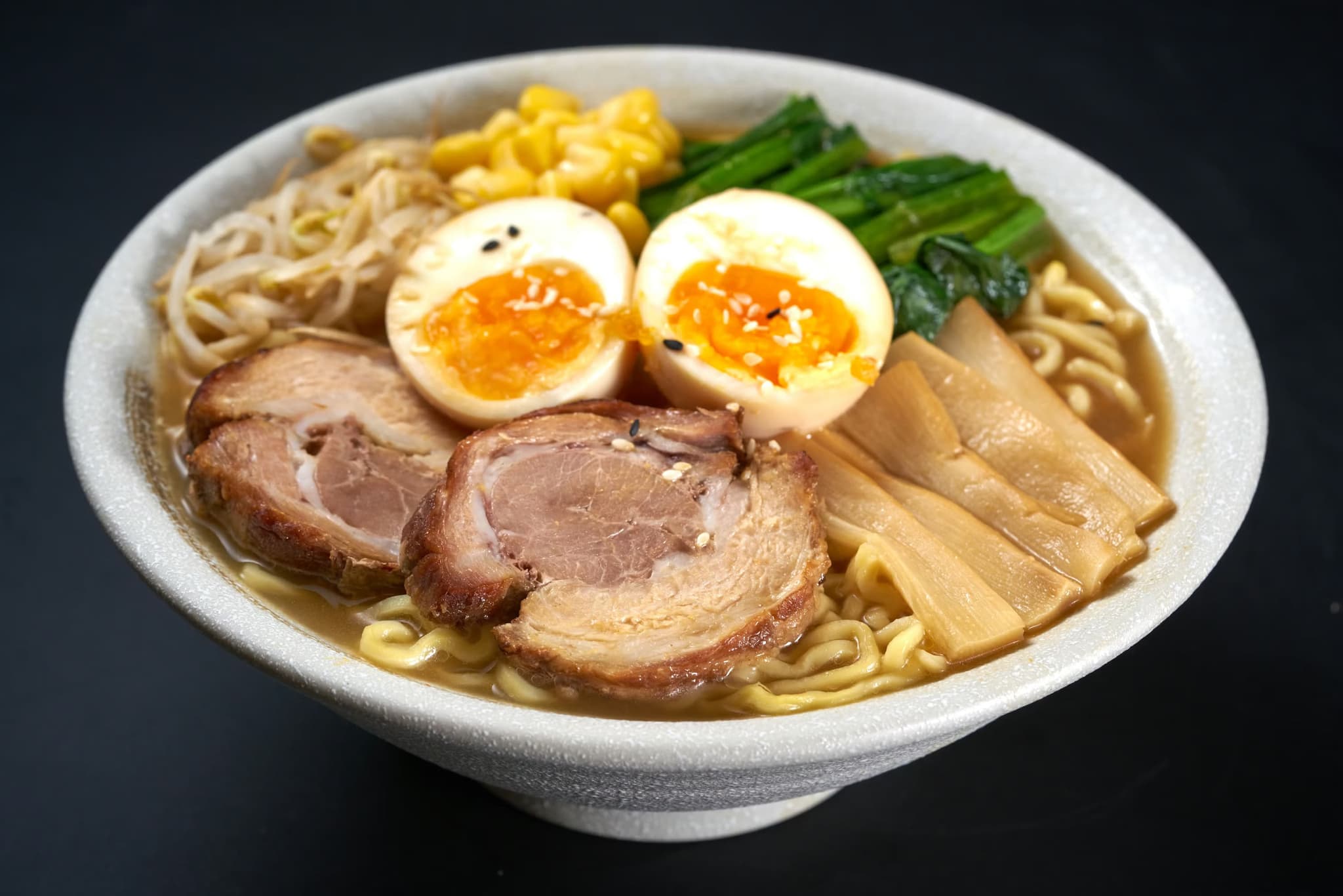 A steaming bowl of ramen with slices of pork, soft-boiled egg halves, bamboo shoots, spinach, bean sprouts, and corn in rich broth.
