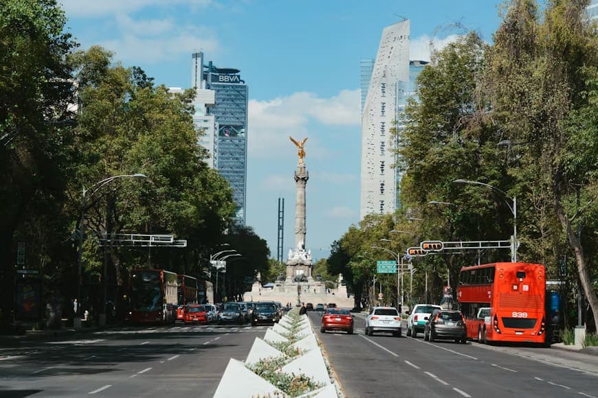 Wide street flanked by tall buildings and trees leads to the Angel of Independence monument. Red buses and cars create an urban bustling scene.