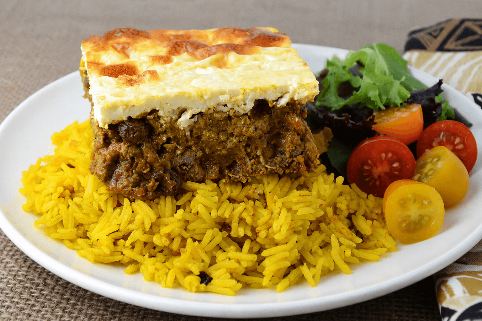 A white plate of bobotie served with yellow rice and tomatoes on the side