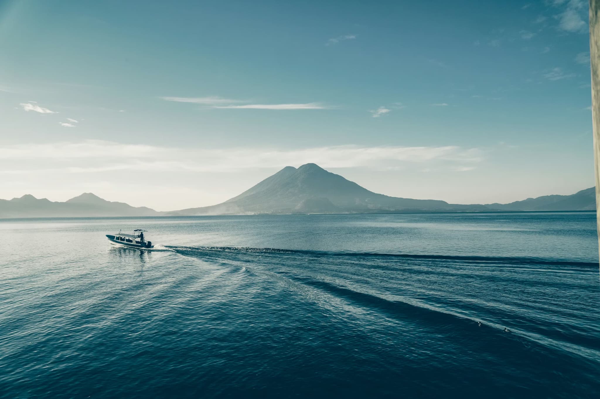A lone boat creates ripples on a vast blue lake with a distant mountain under a clear sky, evoking a tranquil, expansive, and peaceful feeling.