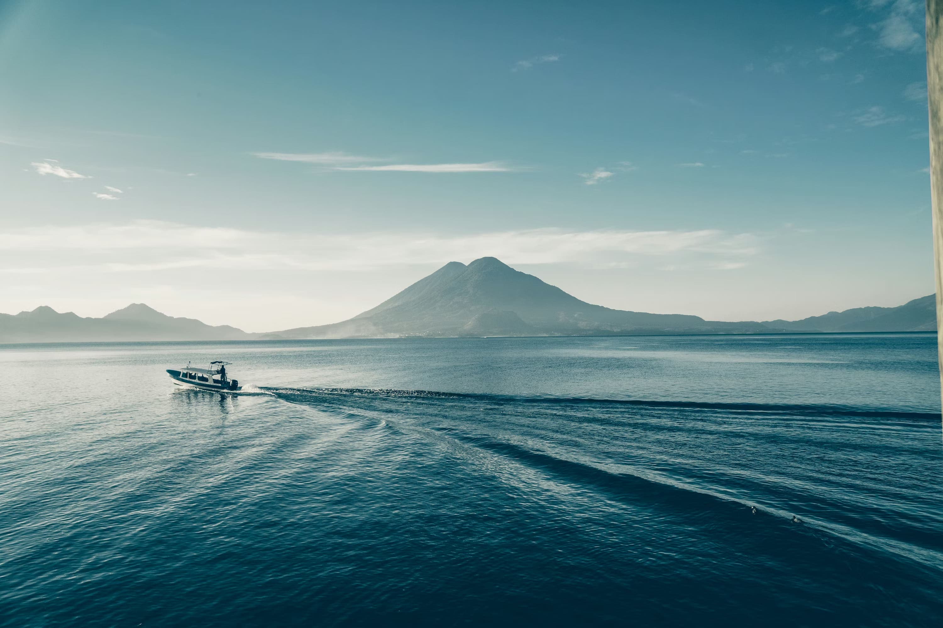 A lone boat creates ripples on a vast blue lake with a distant mountain under a clear sky, evoking a tranquil, expansive, and peaceful feeling.