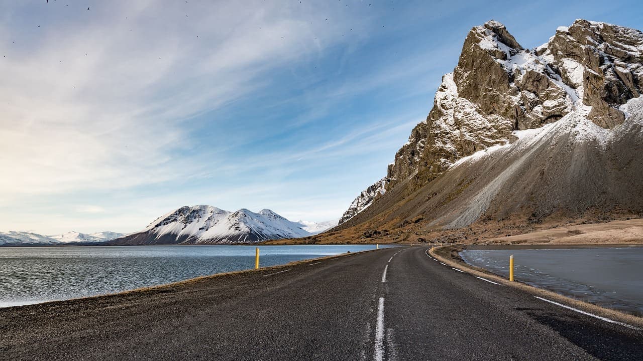 The Iceland Ring Road stretching along a coastal mountain landscape with snow covered peaks and calm sea under a blue sky.