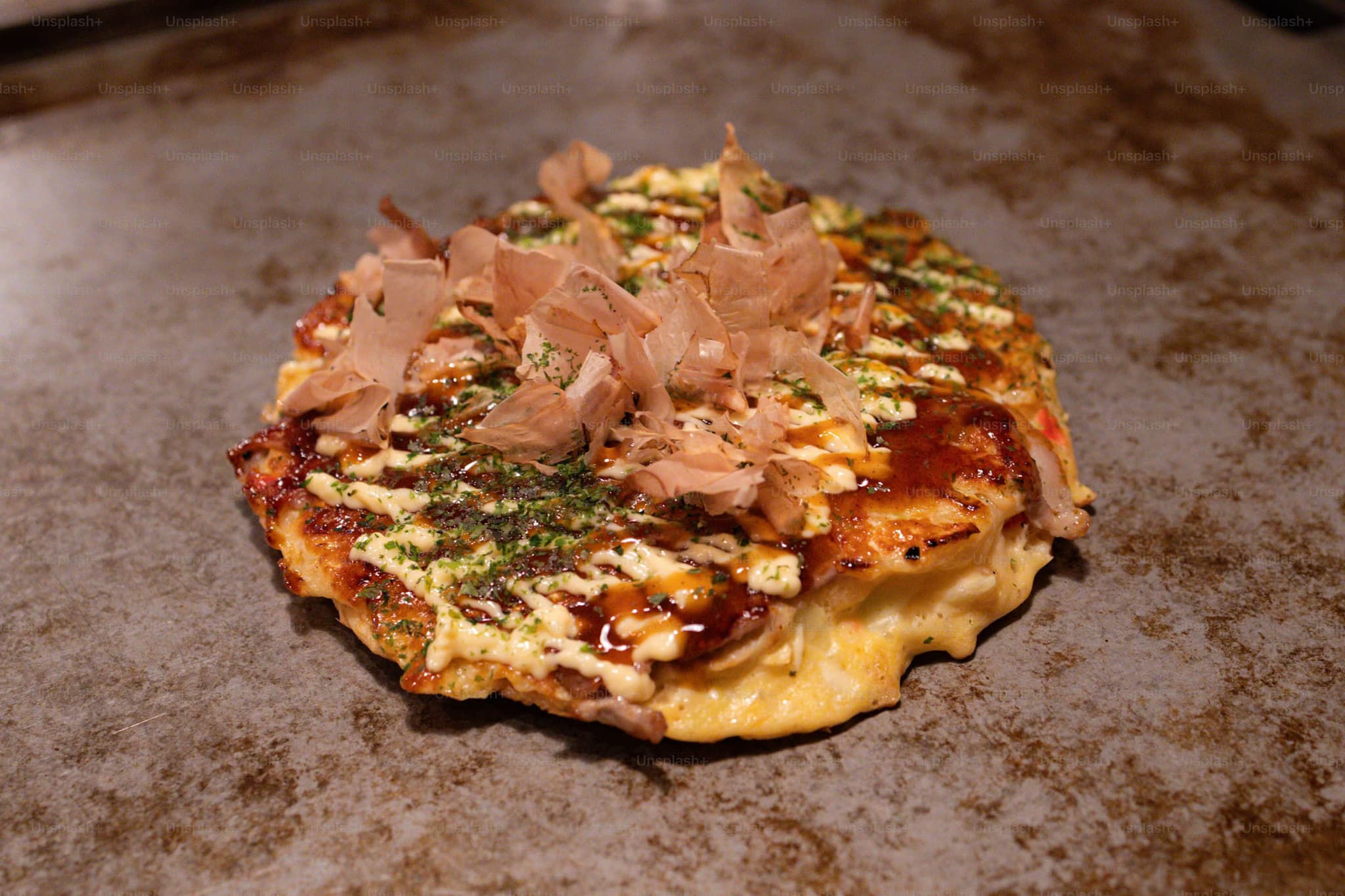 Okonomiyaki pancake on a griddle, topped with bonito flakes, drizzled with mayonnaise and sauce. The dish looks savory and appealing.