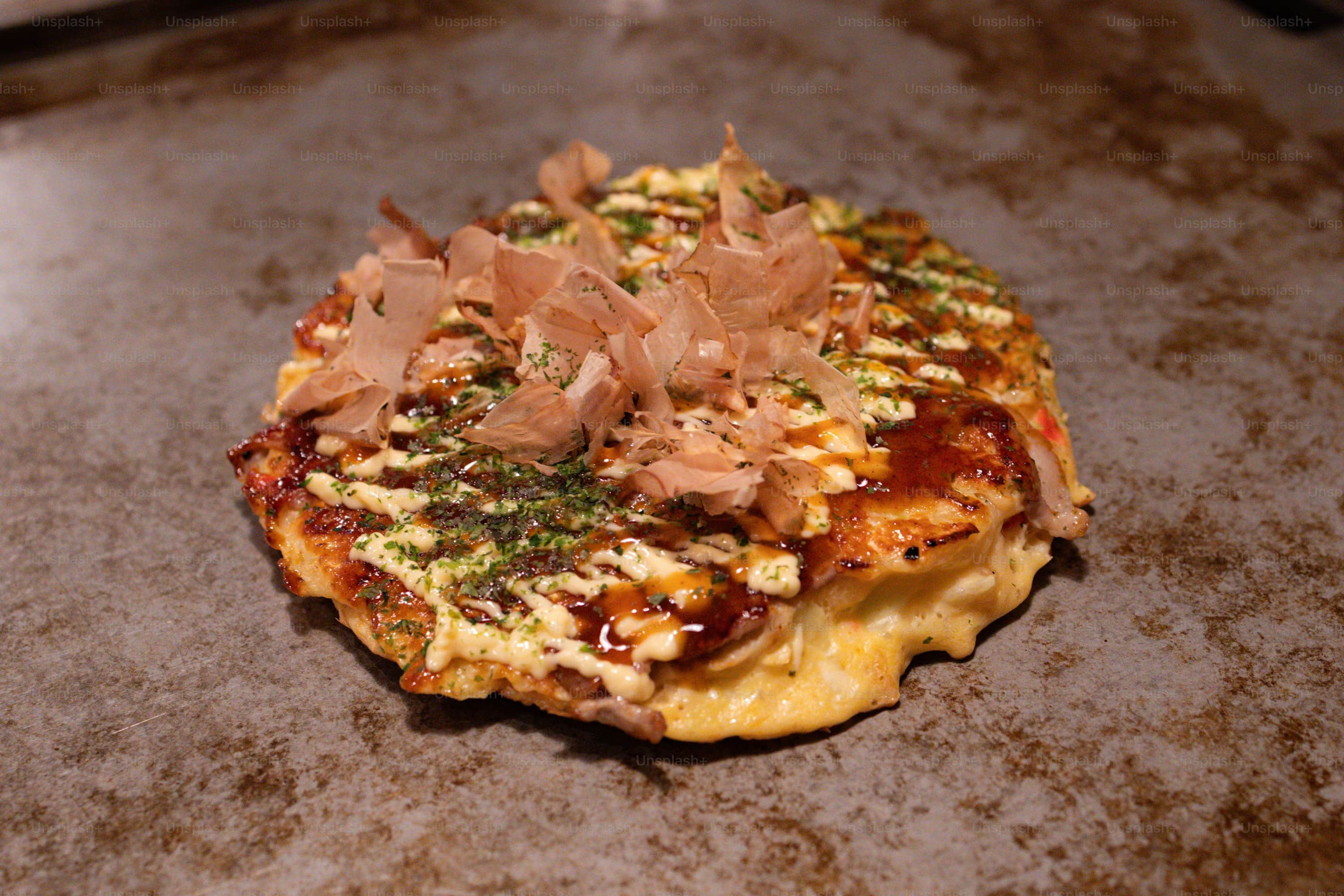 Okonomiyaki pancake on a griddle, topped with bonito flakes, drizzled with mayonnaise and sauce. The dish looks savory and appealing.