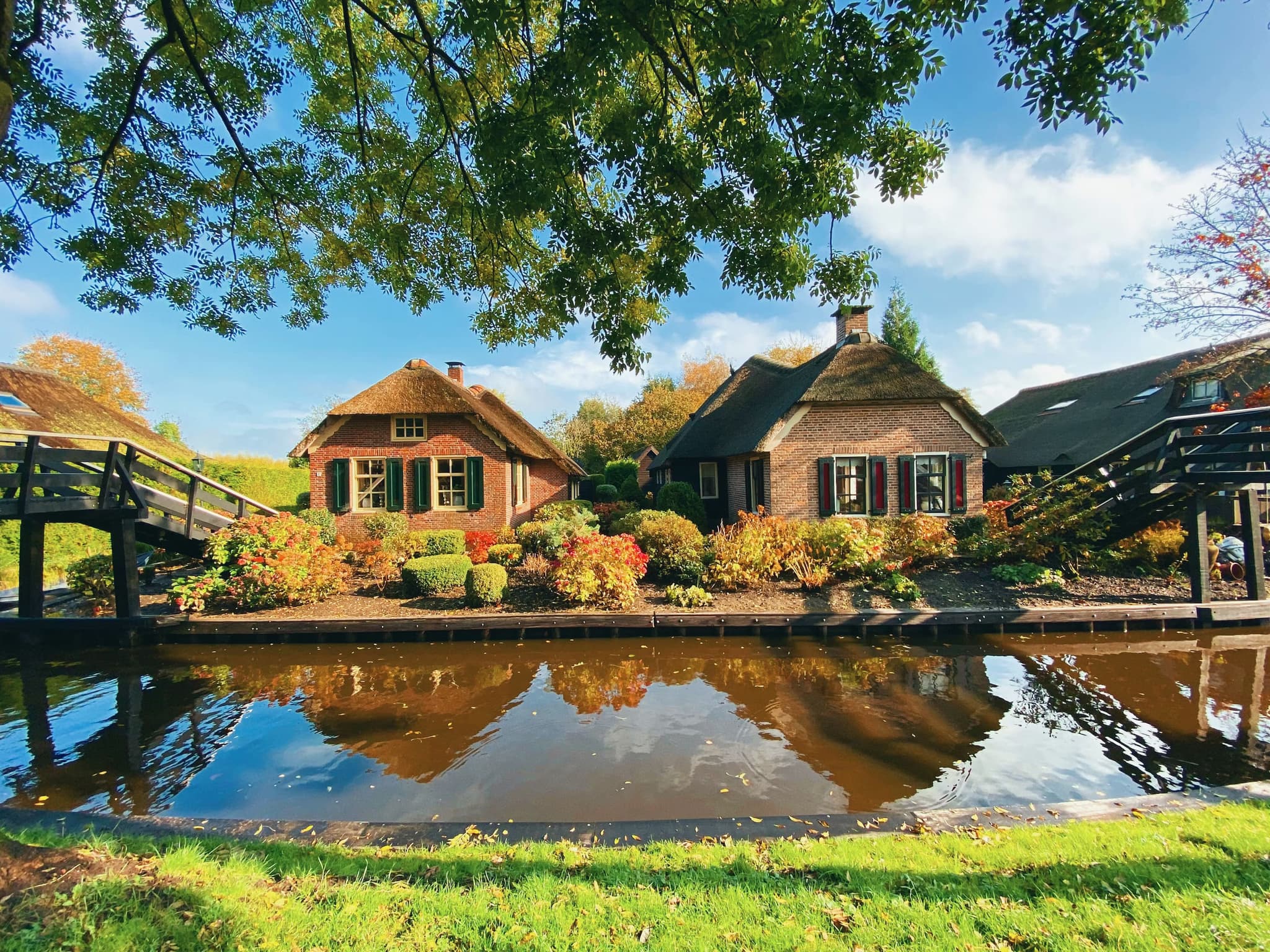Quaint brick cottages with thatched roofs and colorful gardens line a tranquil canal. Wooden bridges connect the lush landscape under a clear blue sky.