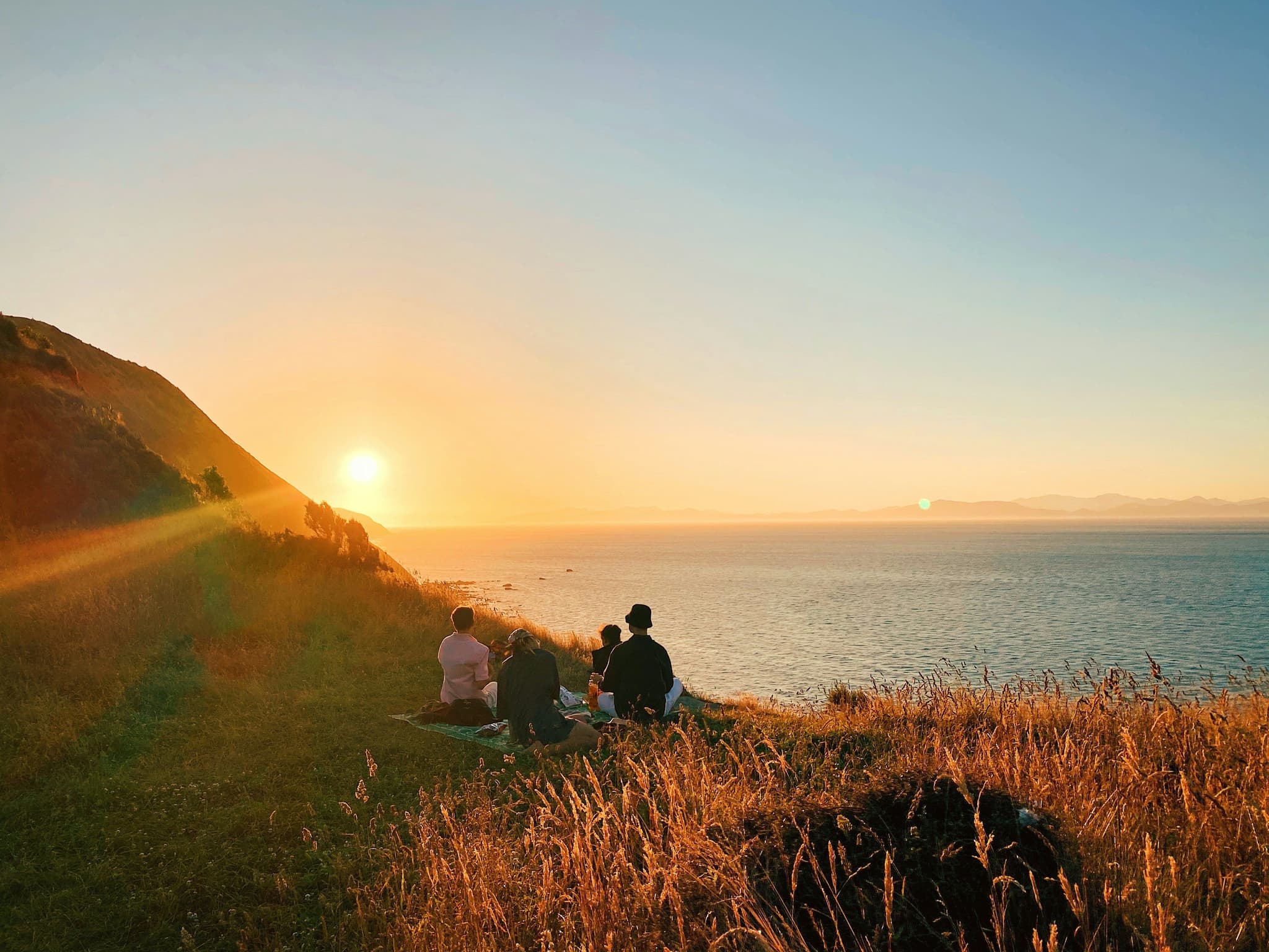 people picknicking on a hill overlooking the ocean during sunset