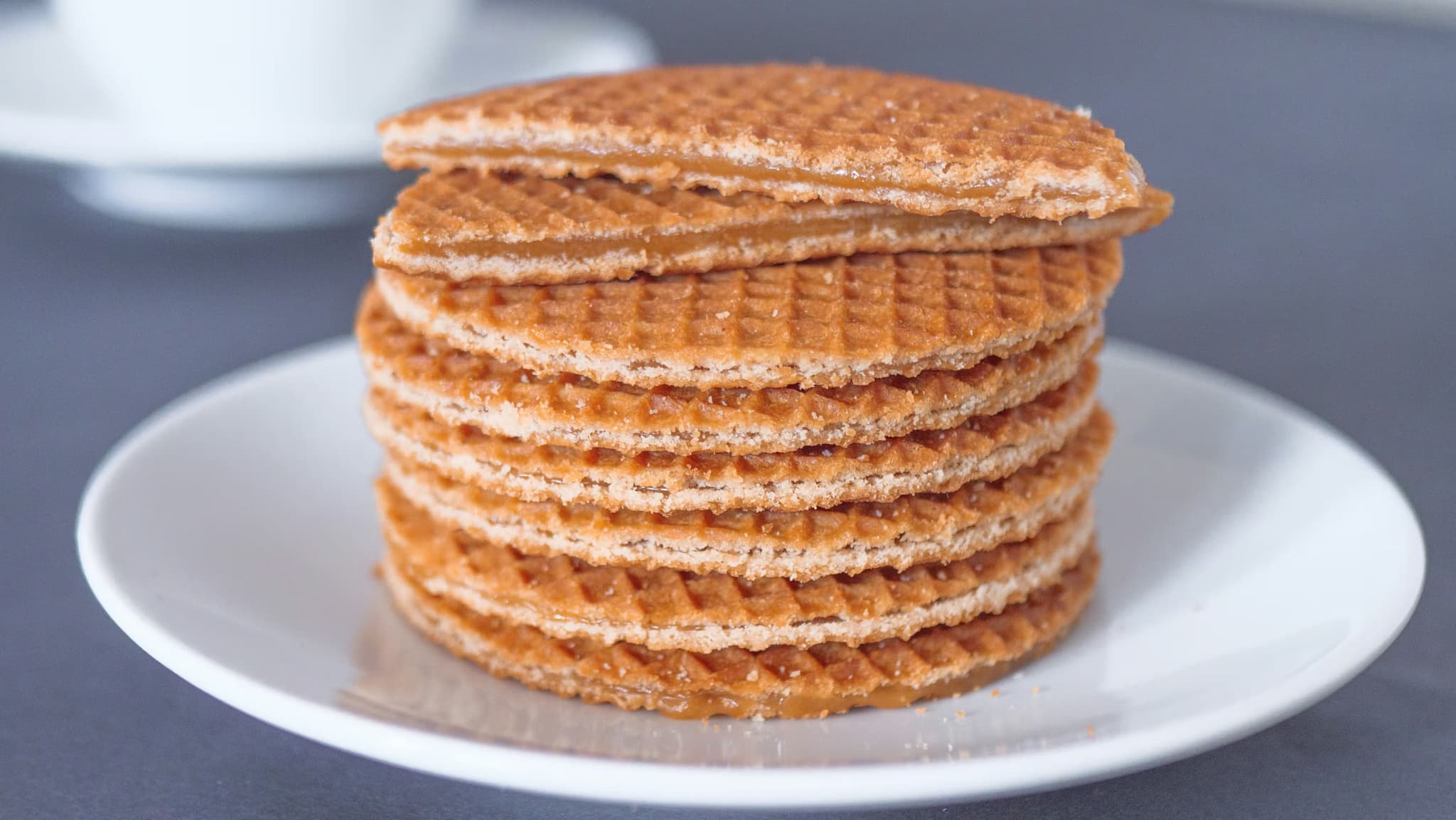 Stack of round, golden-brown stroopwafels on a white plate, with one partially broken on top. Background is softly blurred, creating a cozy, inviting feel.