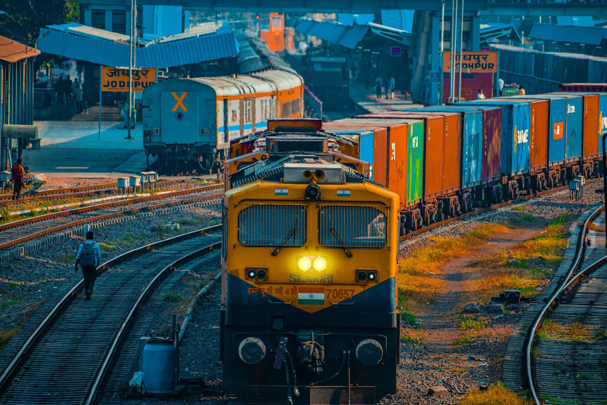 A vibrant train station scene with a yellow freight train in the foreground, colorful cargo containers, and people walking along tracks. The setting is bustling and dynamic.