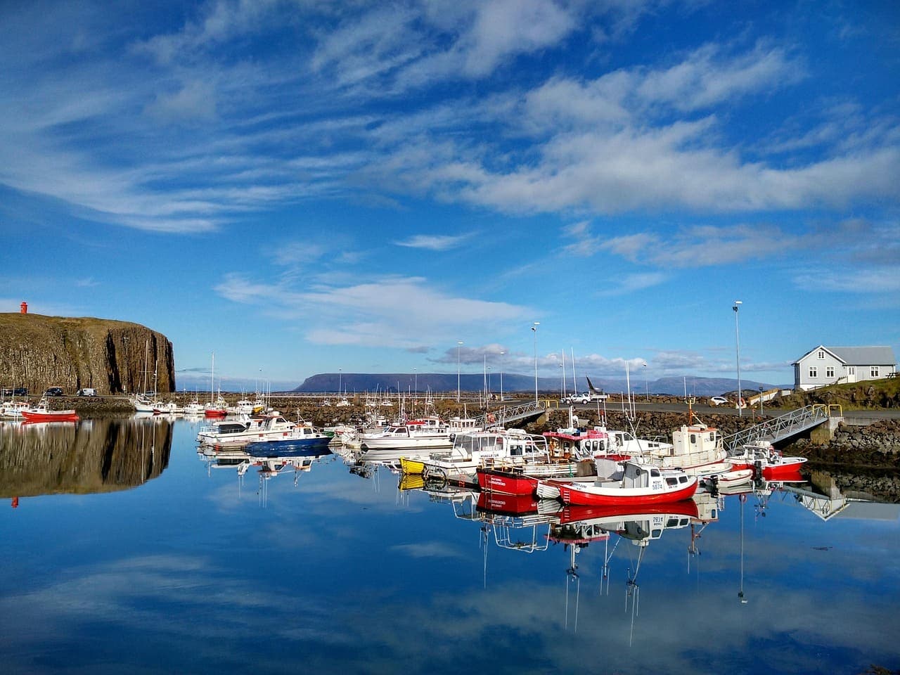 Small fishing harbour with colourful boats reflected in still water under a bright blue sky in West Iceland.
