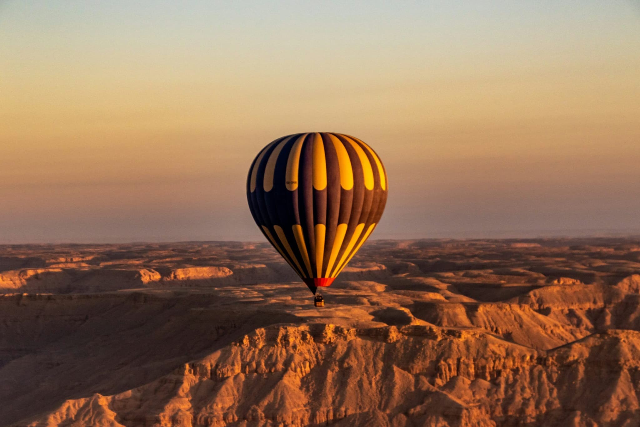 Hot air balloon with yellow and blue stripes floats over a vast desert landscape at sunset, casting long shadows and evoking a sense of tranquility.