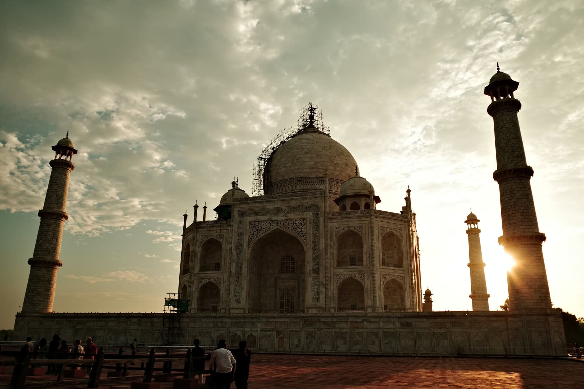 Silhouetted view of the Taj Mahal at sunrise, with the sun peeking between minarets. The sky is partly cloudy, creating a serene and majestic atmosphere.