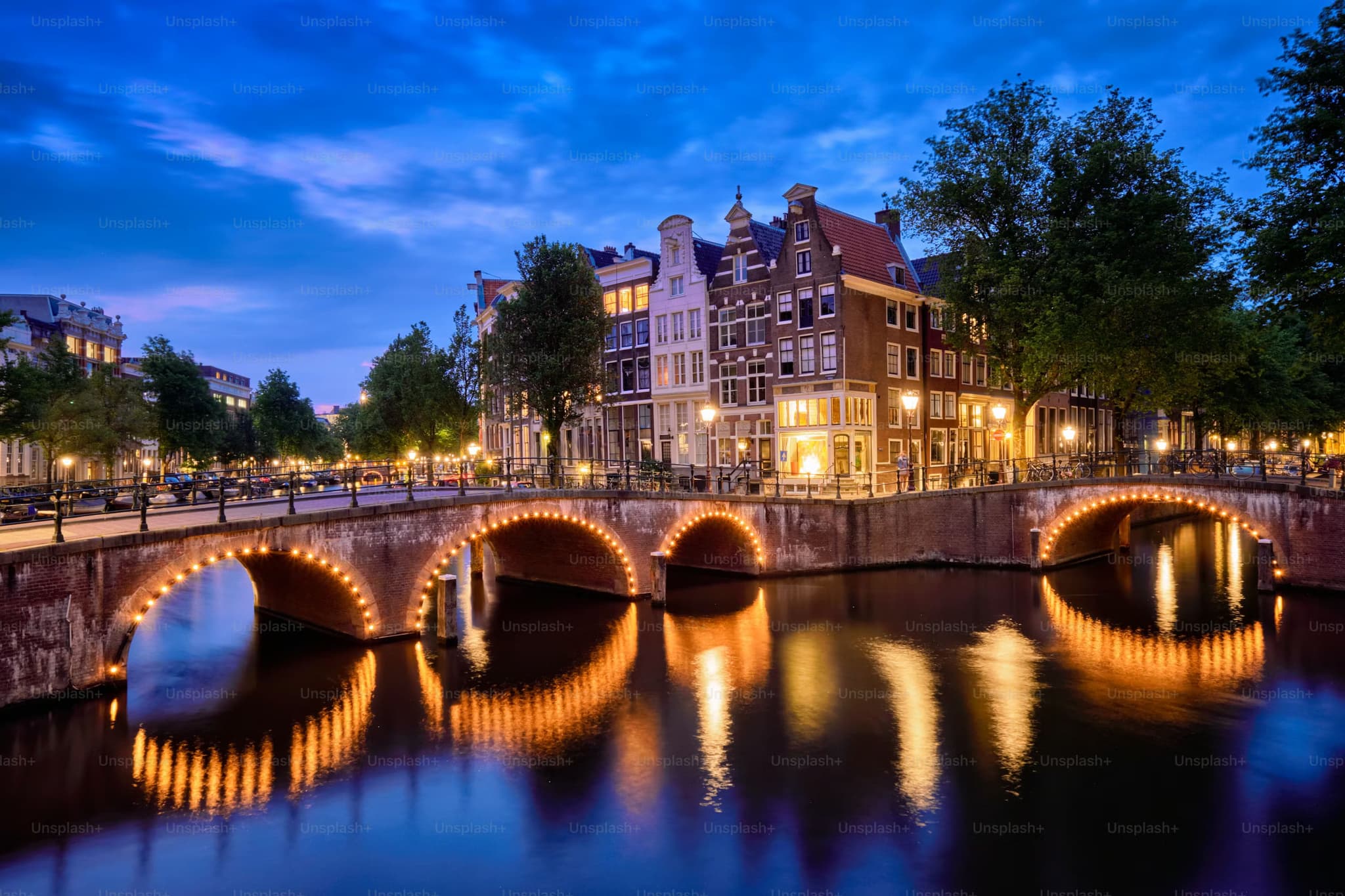 Illuminated bridges over an Amsterdam canal at dusk, with historic buildings and trees reflecting on the water, creating a serene ambiance.