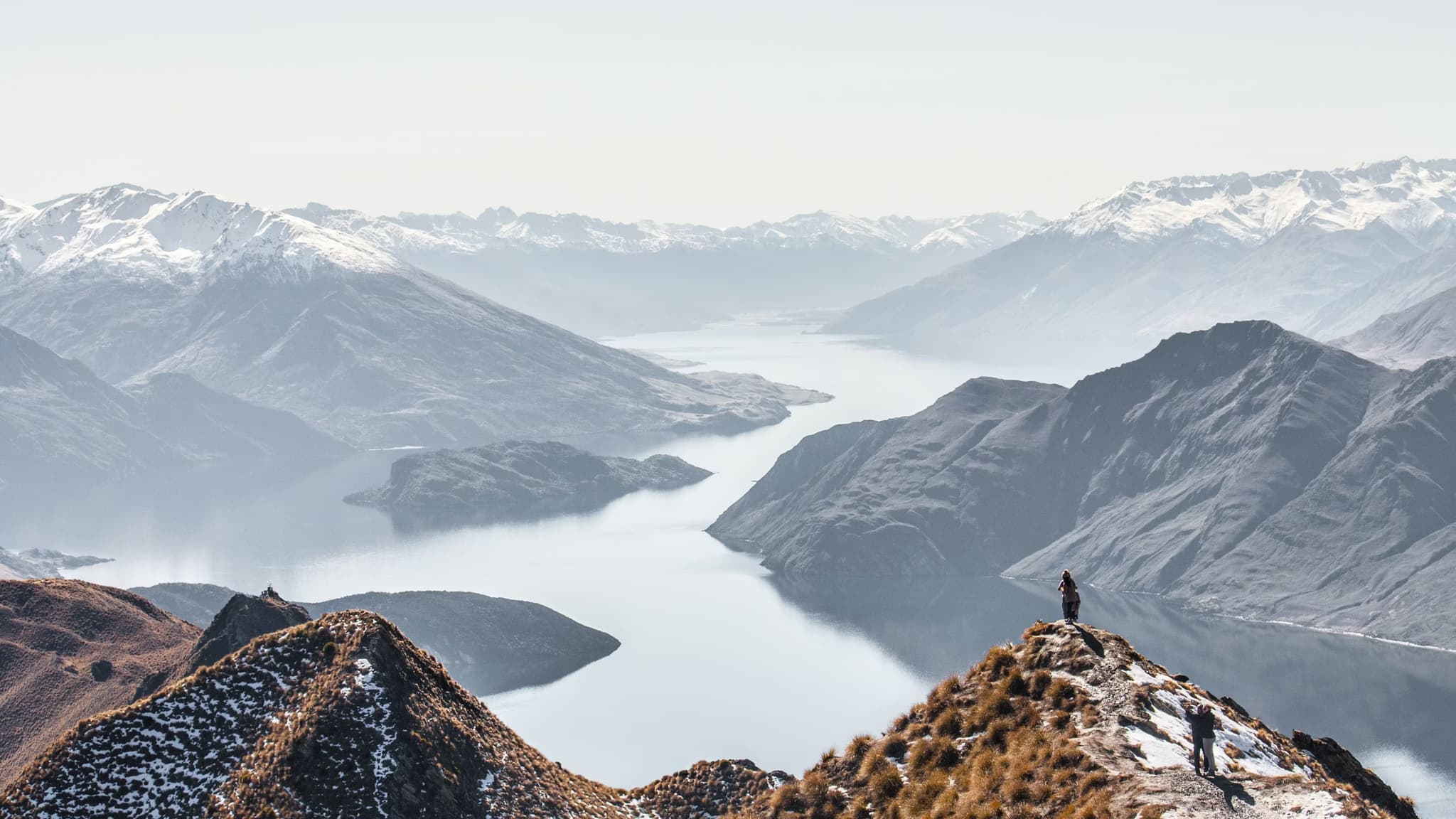 A hiker stands atop a snow-dusted ridge overlooking a vast mountain range and a serene lake. The scene conveys a sense of tranquility and awe.