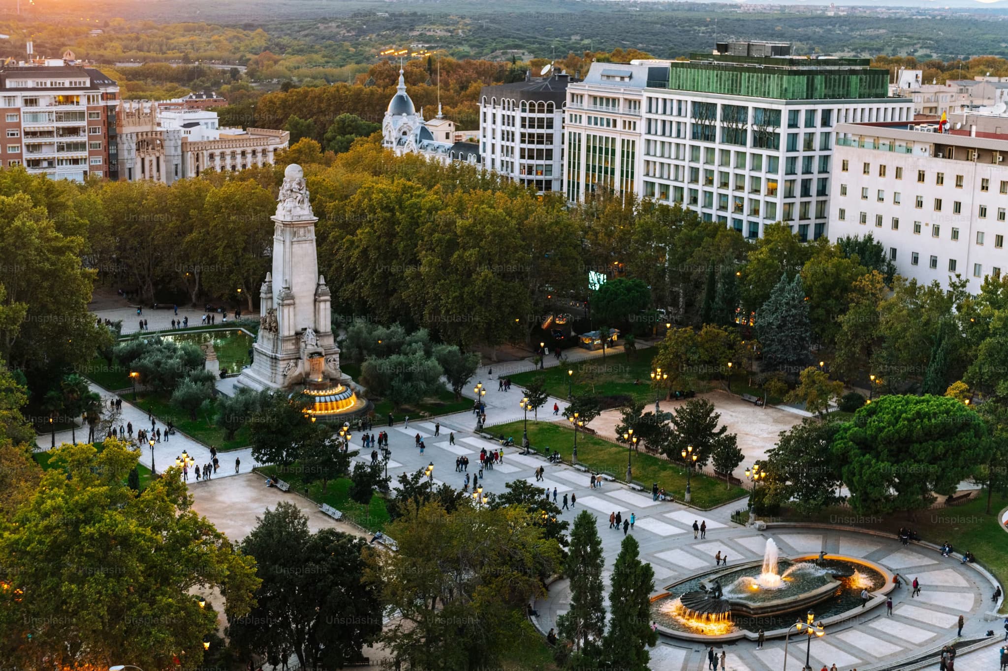 Aerial view of a vibrant city park at dusk featuring a large monument at the center. People stroll along the tree-lined paths, and a lit fountain adds charm. Surrounding high-rises are bathed in a soft, warm light, creating a peaceful urban ambiance.