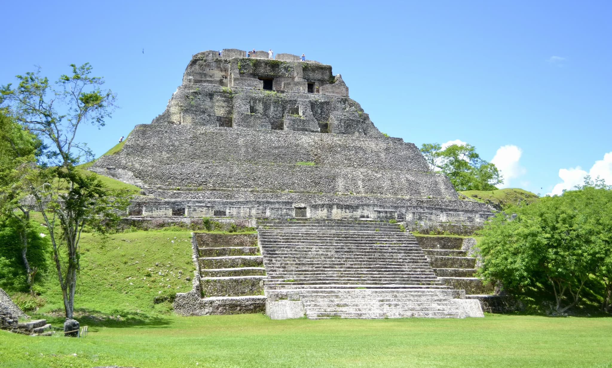 A majestic, ancient stone pyramid with steep, wide steps surrounded by lush greenery under a clear blue sky, evoking a sense of historic grandeur.