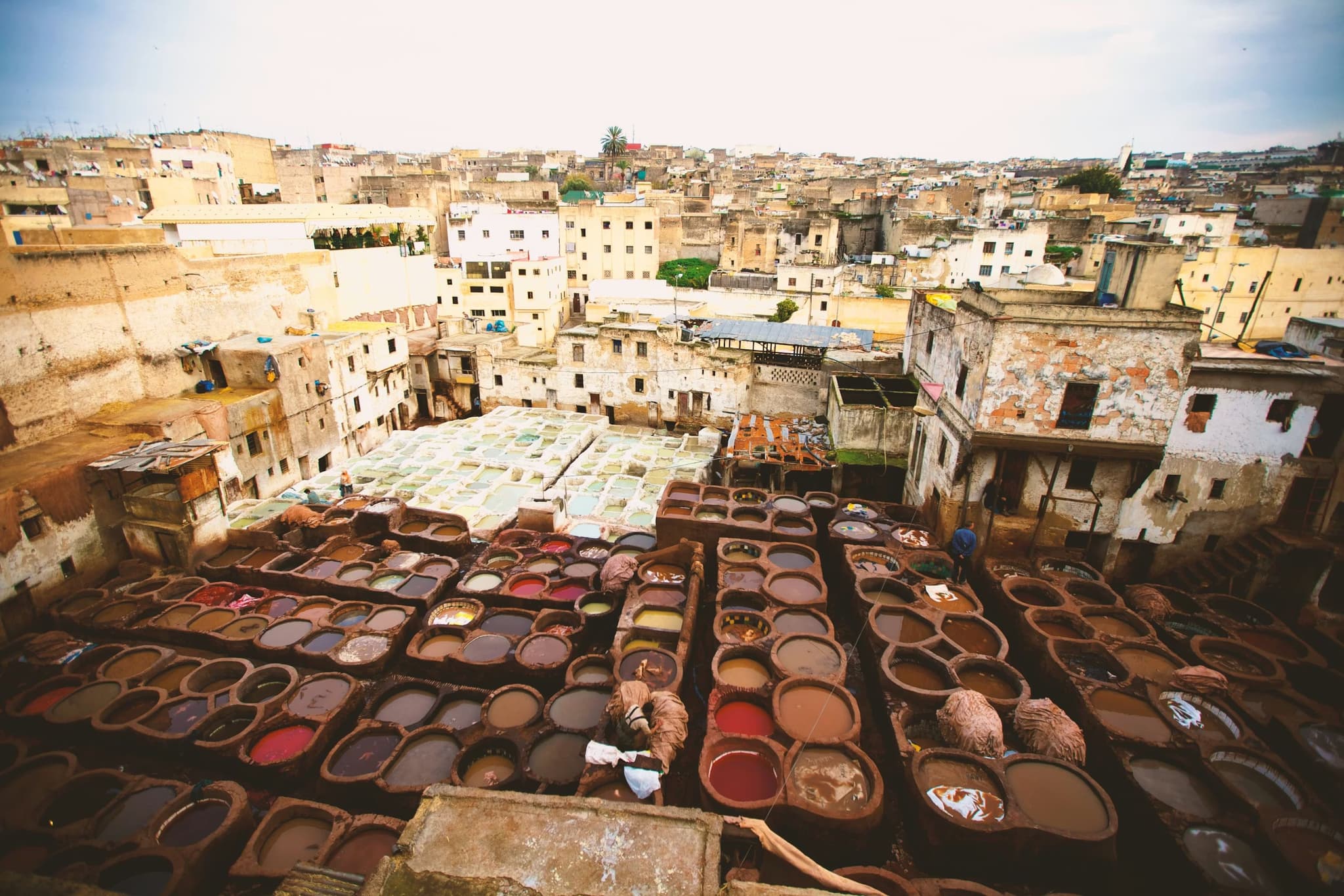 Overlooking a historic tannery in Fez, Morocco, with rows of circular dye pits in earthy tones, surrounded by aged buildings under a cloudy sky.