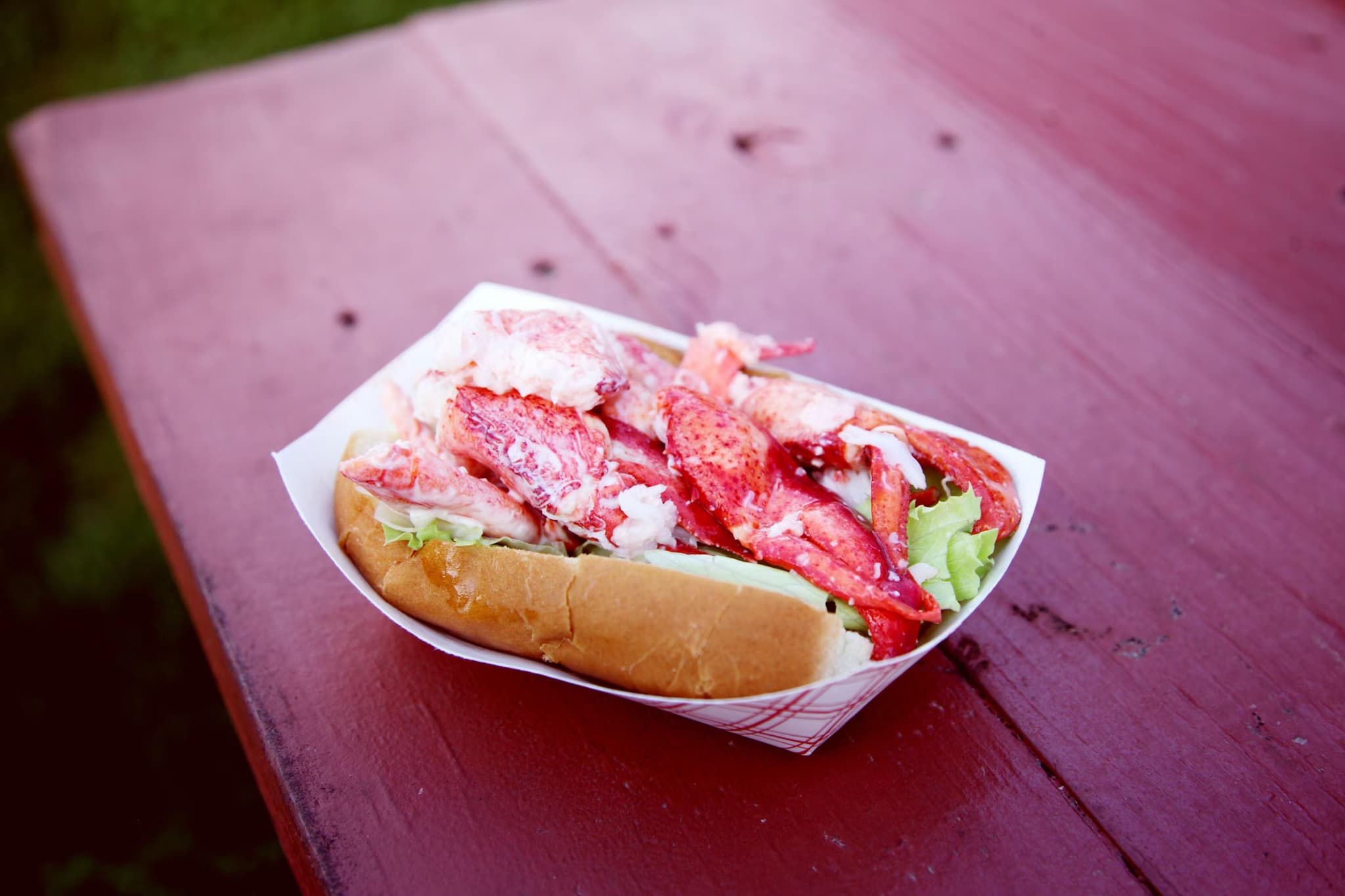 A vibrant lobster roll with chunky lobster meat and lettuce in a toasted bun, served in a paper tray, rests on a rustic red picnic table.