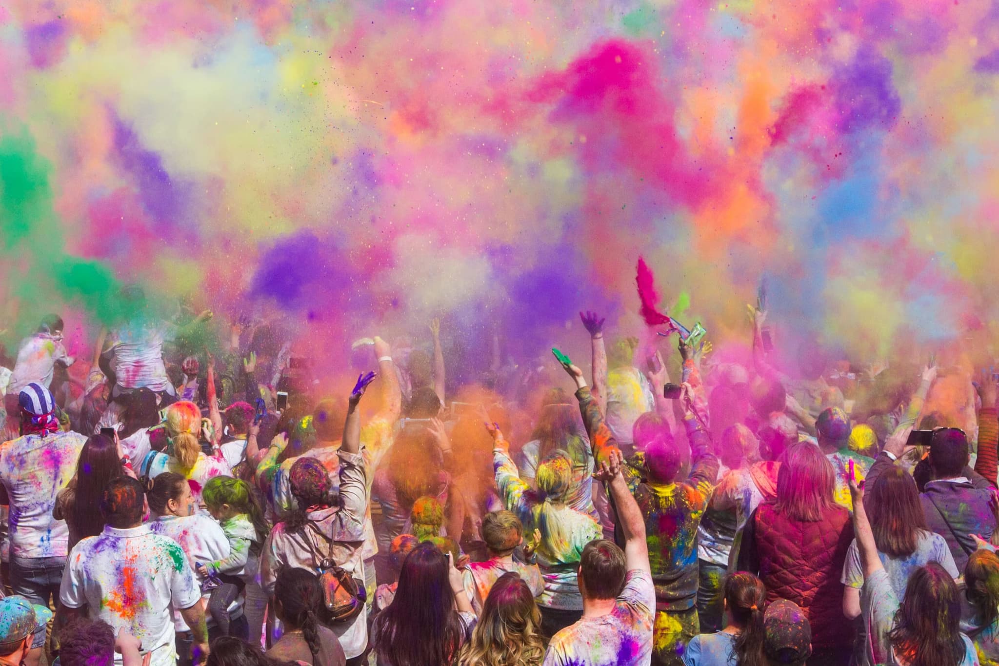 A joyful crowd celebrates by throwing vibrant colored powders into the air, creating an energetic scene filled with pink, yellow, blue, and orange hues.