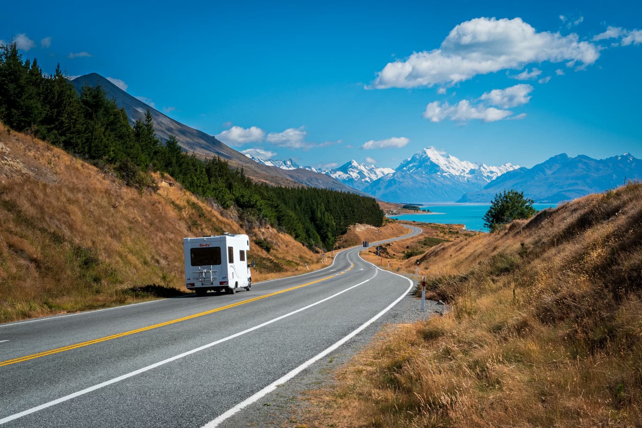A motorhome travels on a winding road through a mountainous landscape. Snow-capped peaks and a turquoise lake are visible under a bright blue sky.