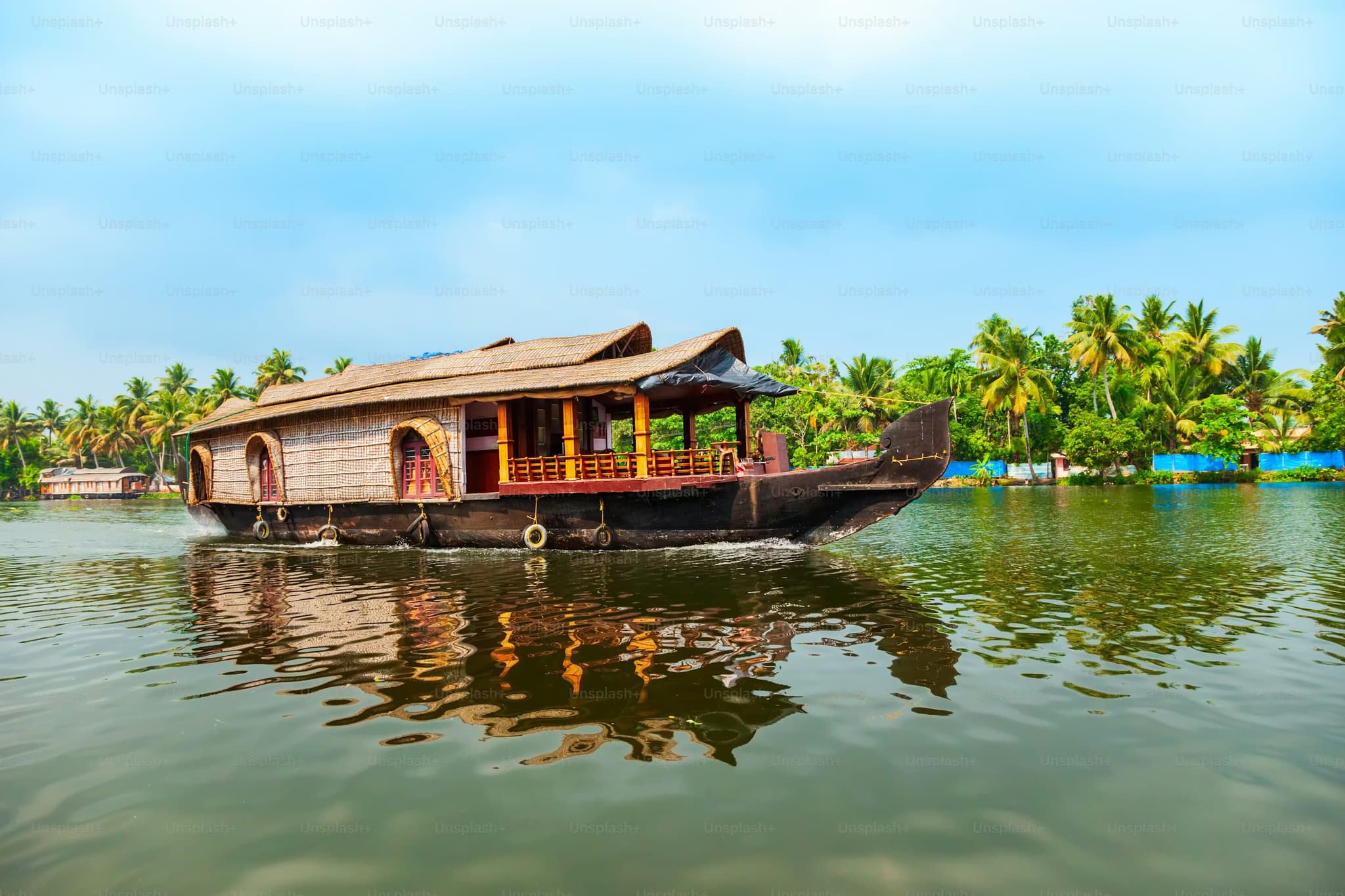 A traditional houseboat floats on a calm river surrounded by lush palm trees under a clear blue sky, creating a serene and picturesque setting.