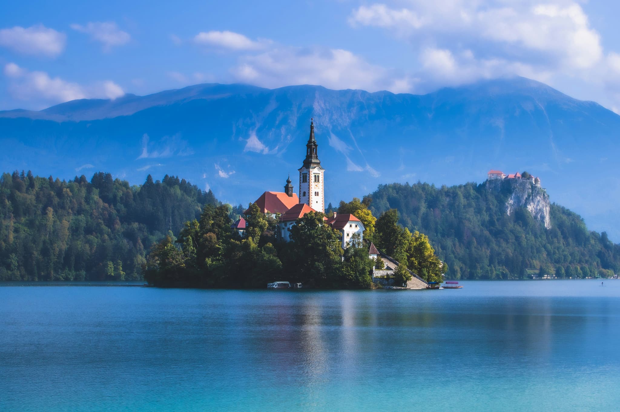 Wide open blue Lake Bled with island with castle tower and mountain backdrop.