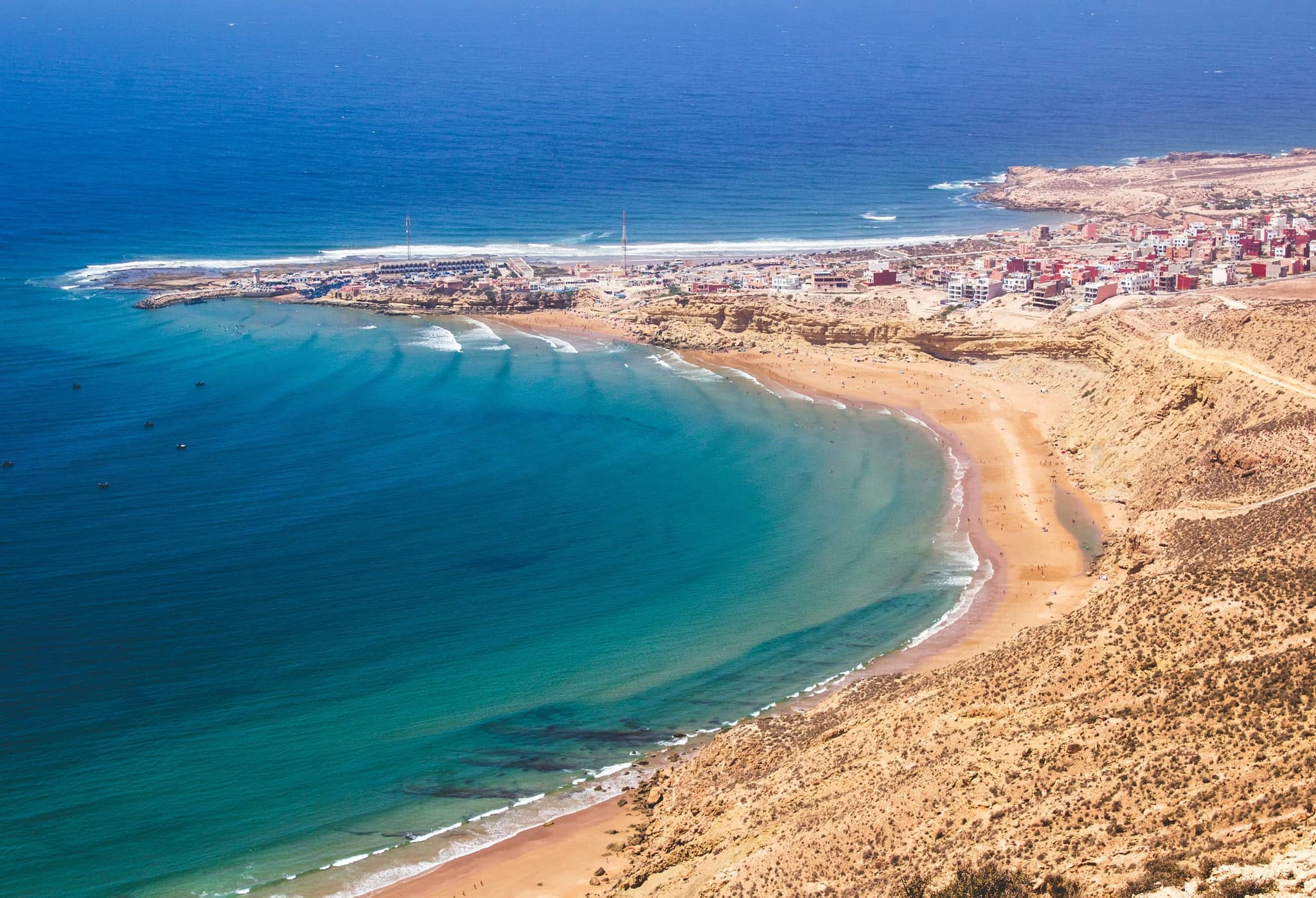 Aerial view of a serene coastal town with turquoise waters and a sweeping sandy beach. Buildings line the shore, creating a peaceful, picturesque scene.