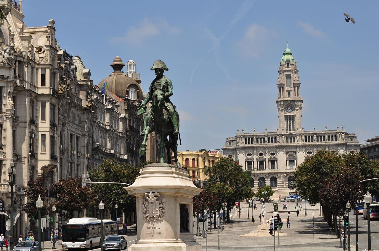 A city square in Porto with historic buildings, a statue in the centre and people walking on a sunny winter day.