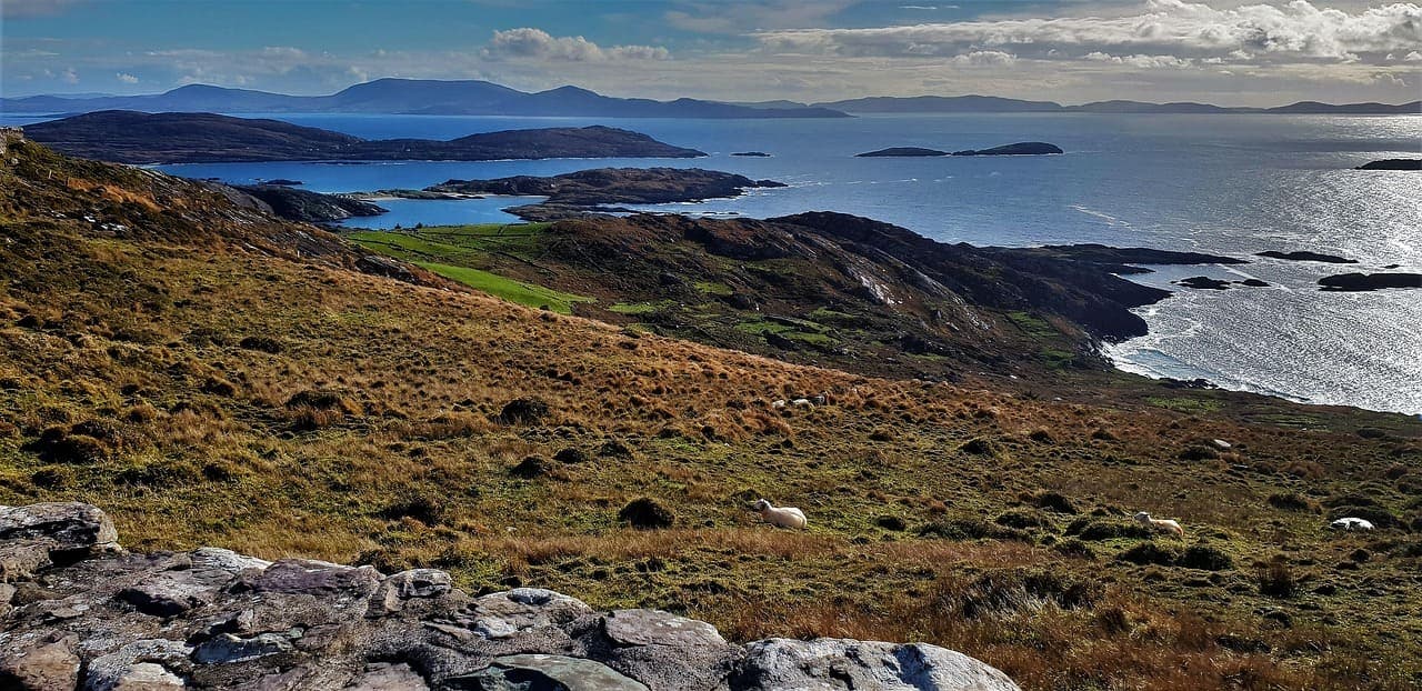 Coastal landscape in southwest Ireland with rolling hills, rocky shoreline and blue ocean views under a bright sky.