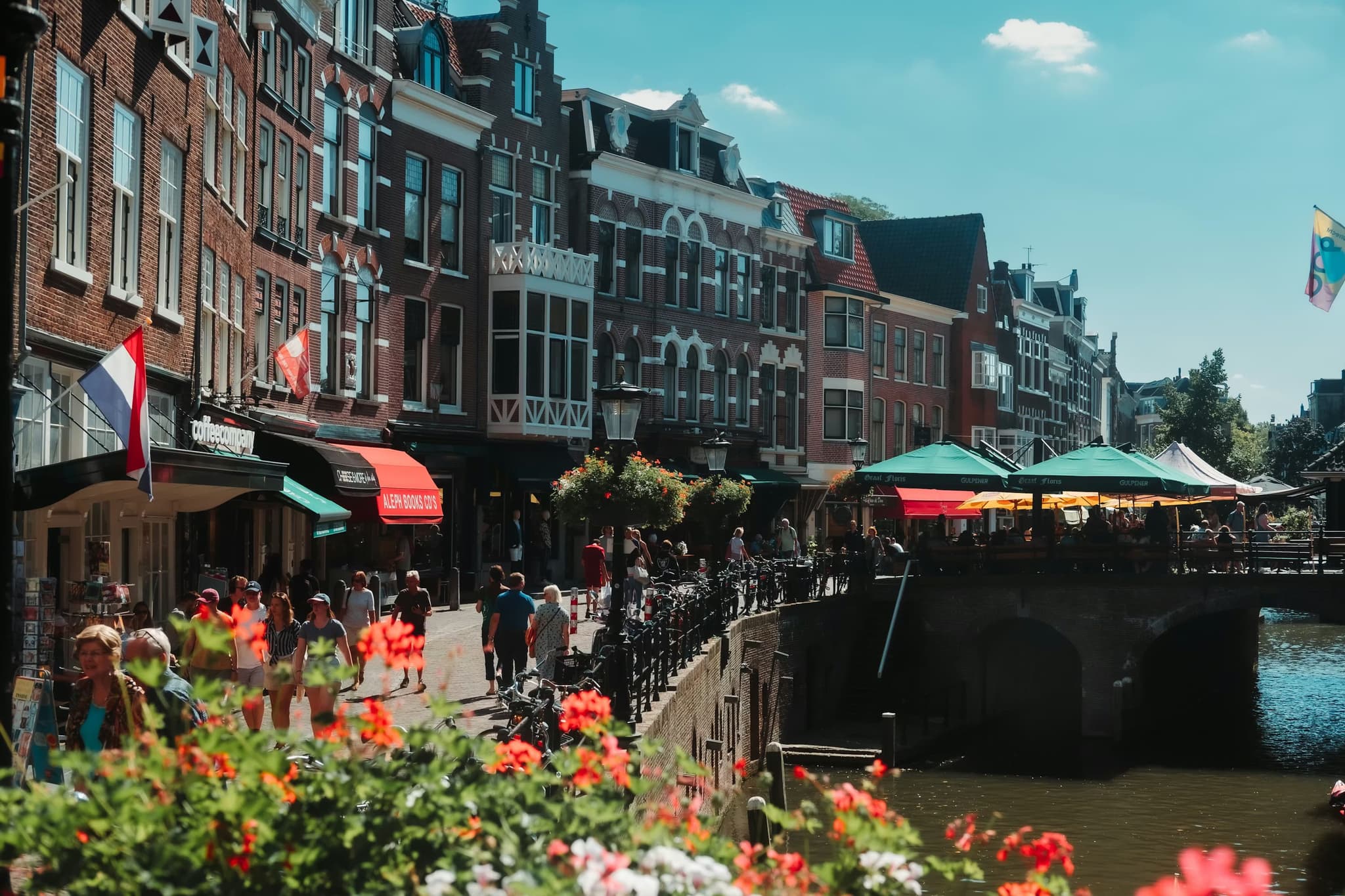 A sunny street in the Netherlands with historic brick buildings, shops, and a canal. People walk and sit at colorful outdoor cafes. Flowers in the foreground.