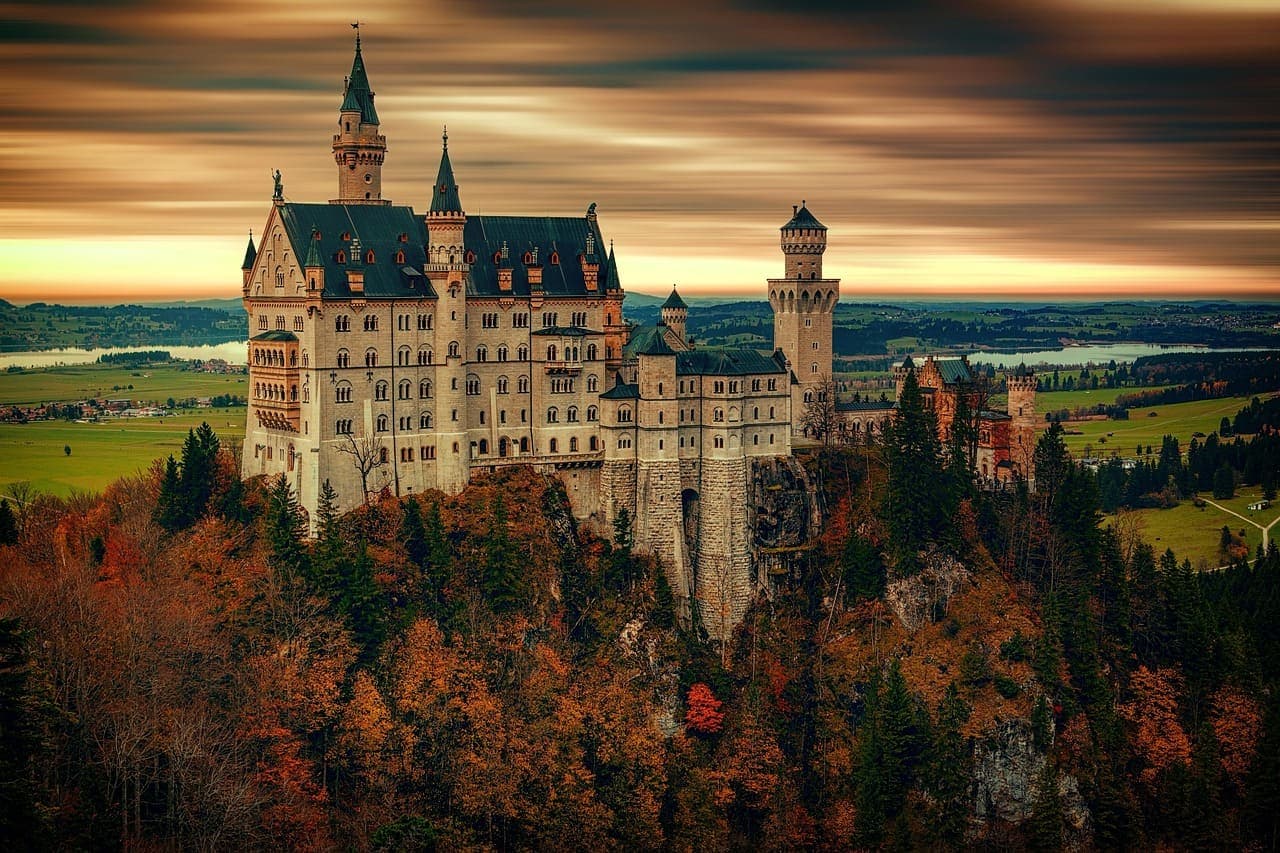 Neuschwanstein Castle on a hill surrounded by autumn coloured trees and rolling countryside under a warm sky.