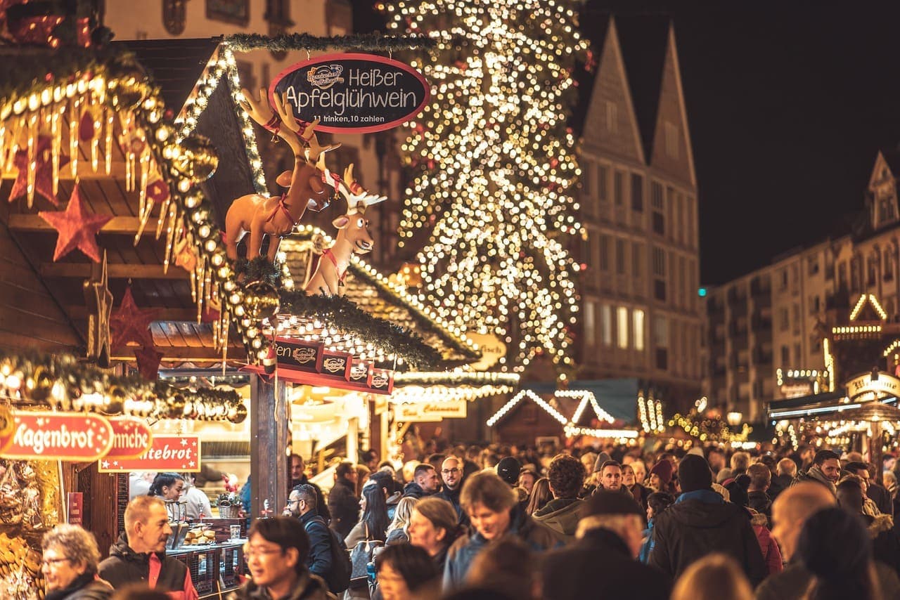 A busy Christmas market at night in Germany with wooden stalls, festive lights and visitors holding hot drinks.